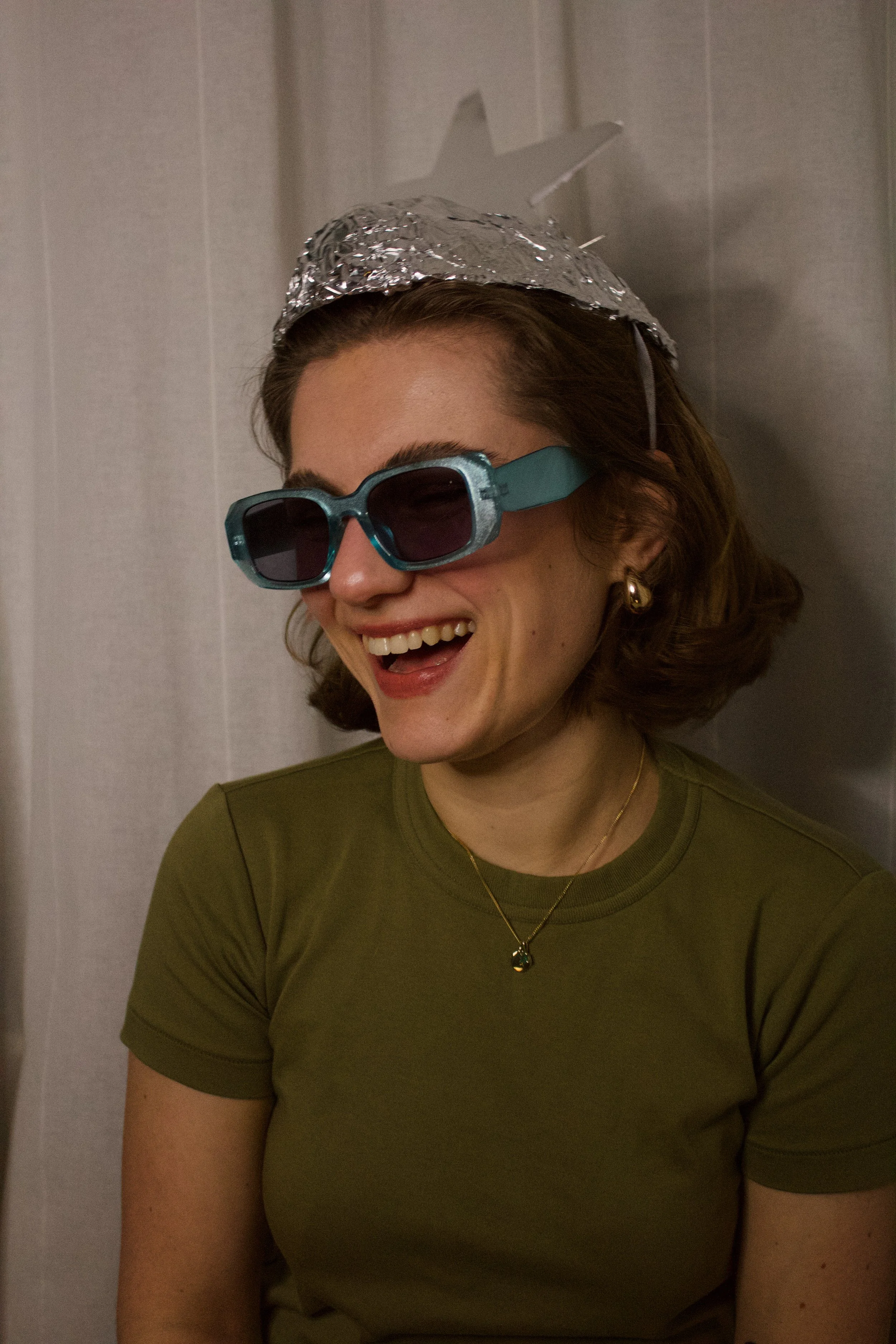 A woman with short brown hair wearing dark sunglasses and a green shirt, smiling and looking to her left, with a silver foil hat on her head.