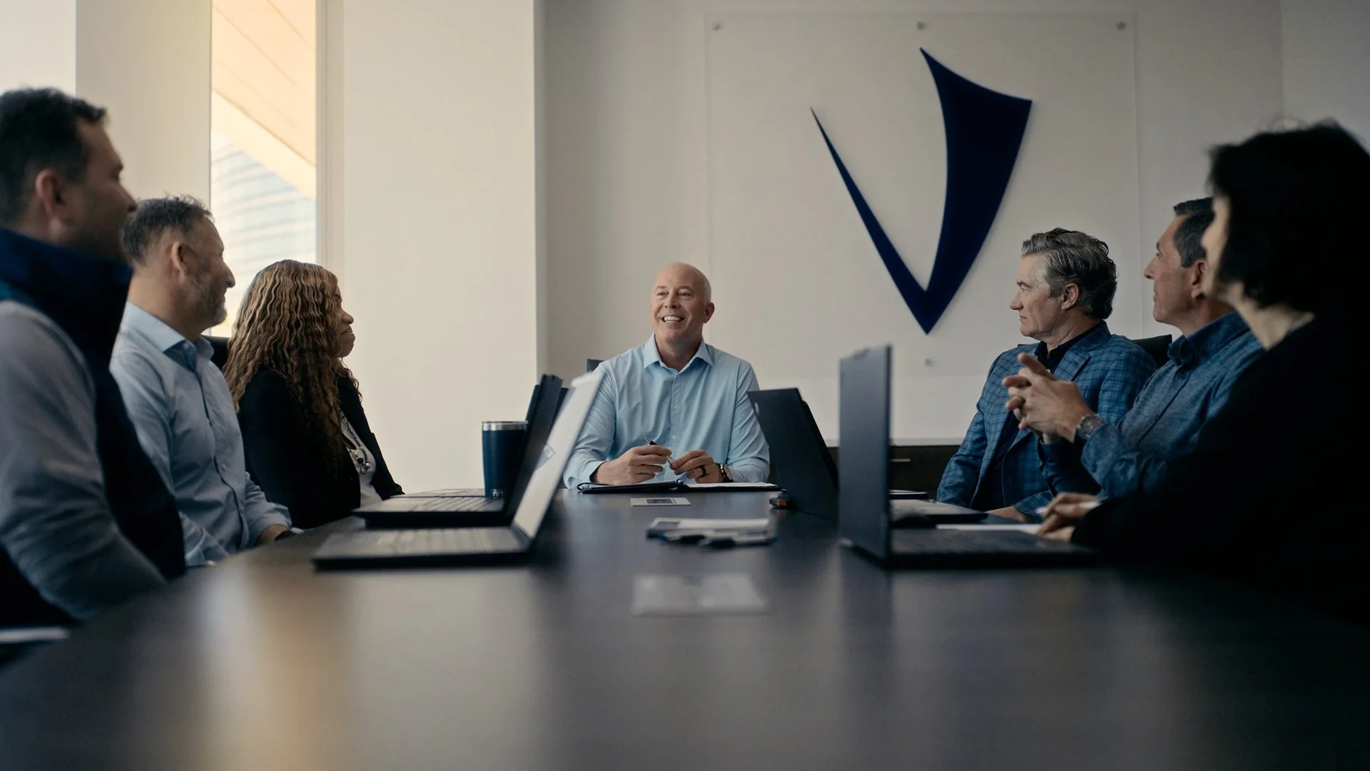 A group of seven professionals sitting around a conference table with laptops, listening to a smiling man in a light shirt leading a meeting in a modern office with a large logo on the wall.