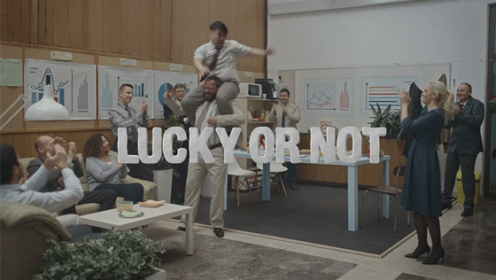 Office party with colleagues, some jumping on a table, others taking photos, with a large sign reading 'LUCKY OR NOT' in the room.