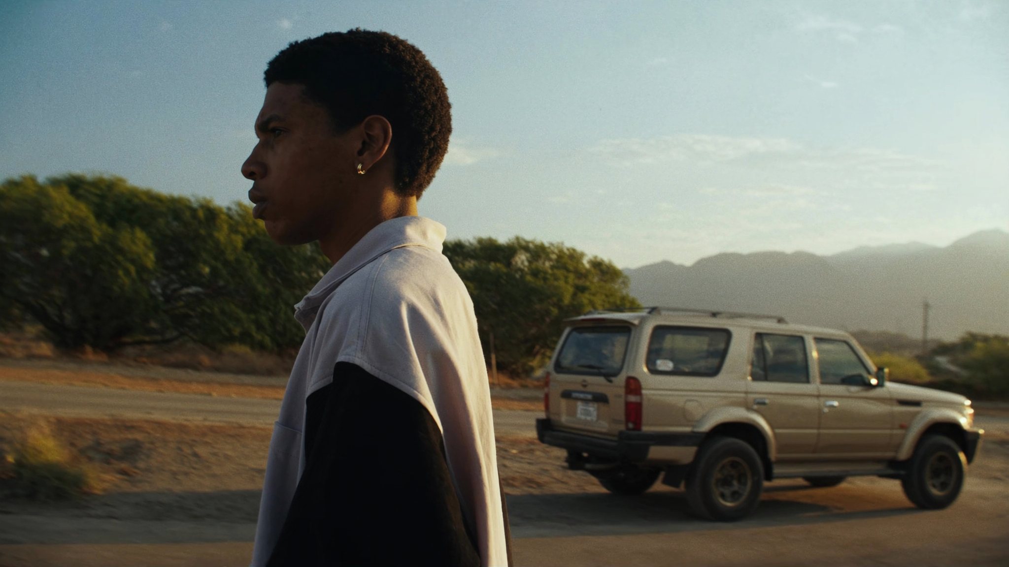 A person with short curly hair and earrings walking along a road with an SUV driving past, trees, and mountains in the background during sunset.