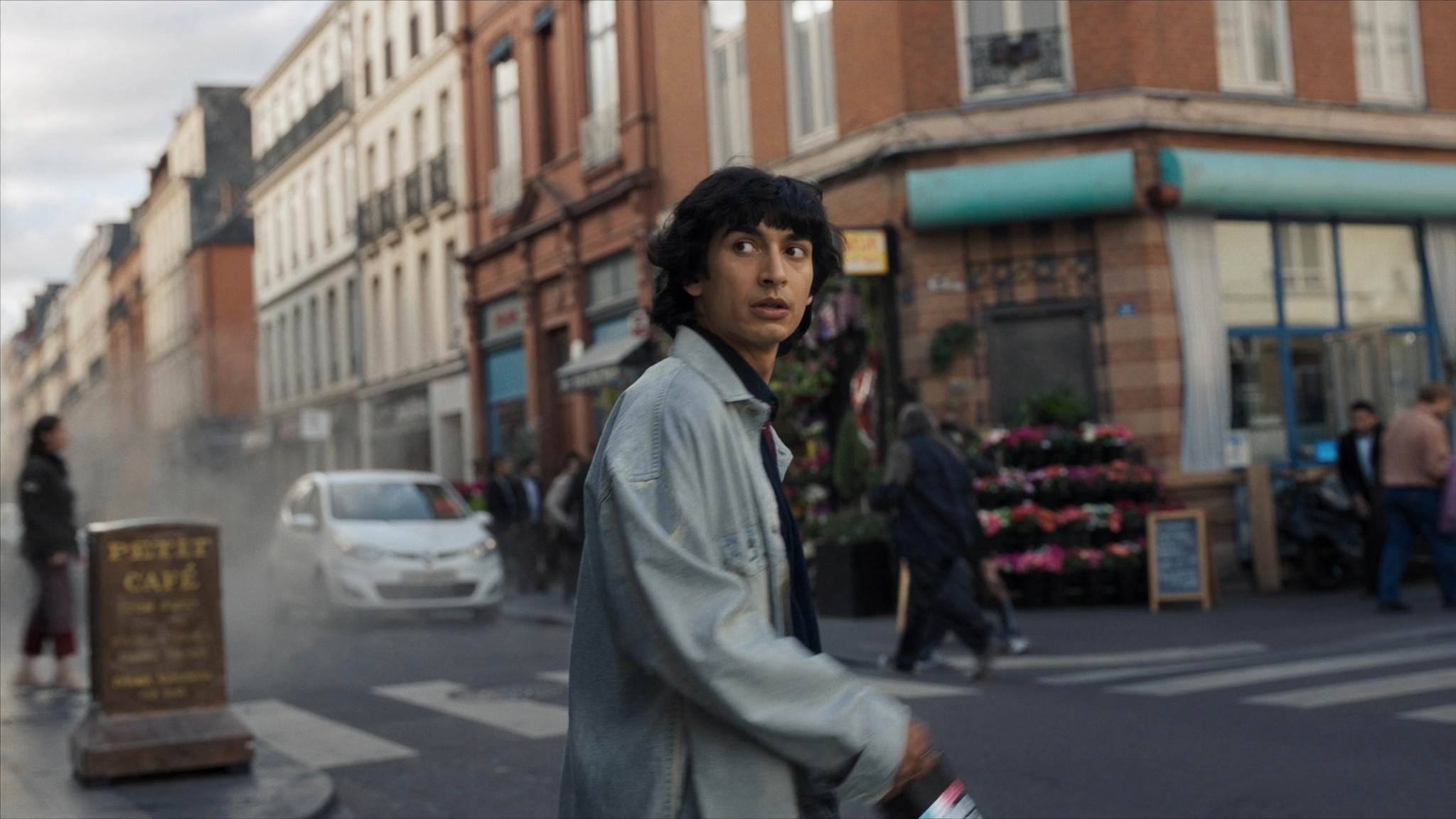 A young man with dark, curly hair looking to the side while walking down a busy city street. There are pedestrians, parked cars, and a flower shop in the background, with a row of older brick buildings lining the street.