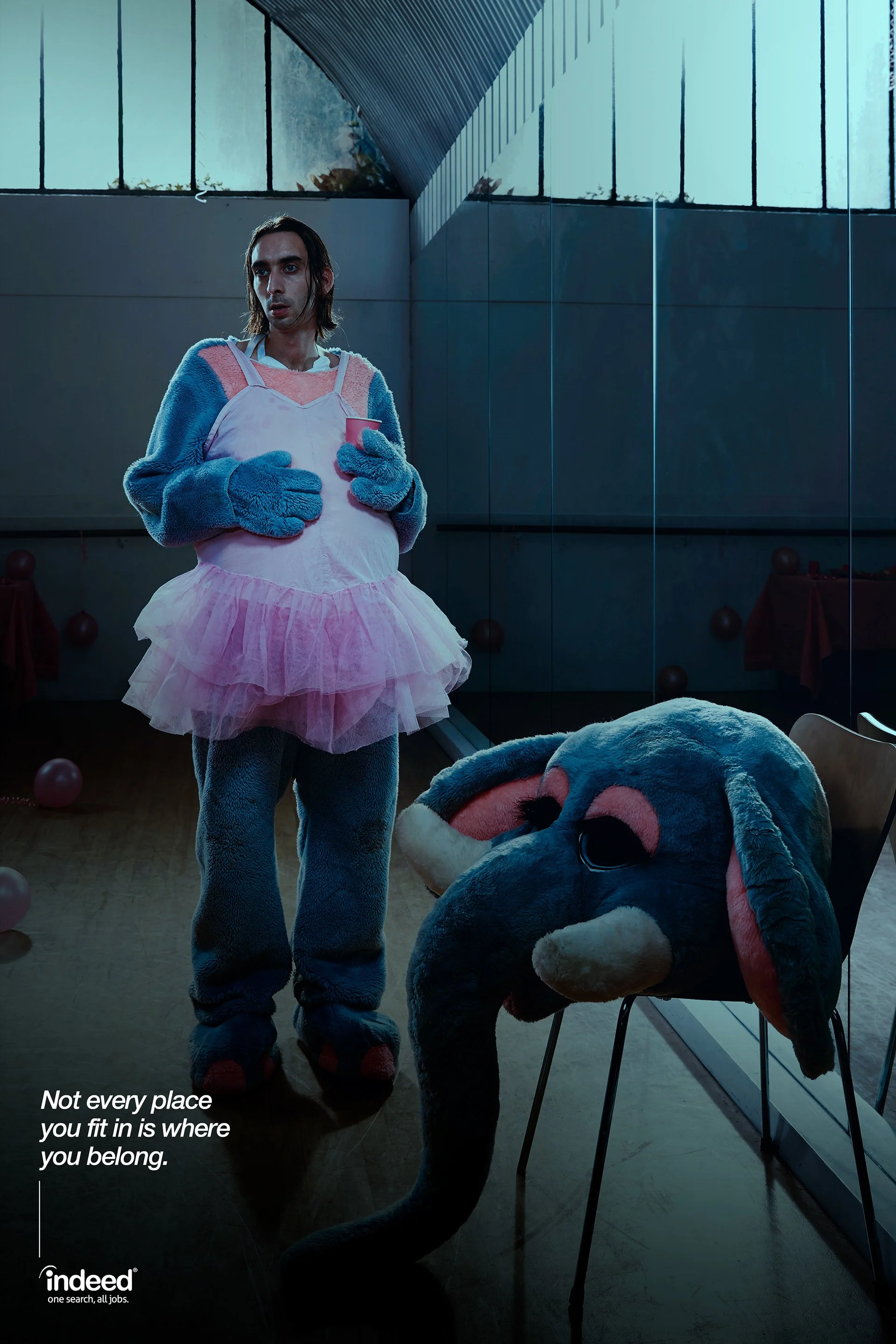 A person dressed in a pink tutu, blue furry gloves, and monster costume paws holding a pink cup, standing indoors near a reflective wall. The reflection shows the person's face. There is a large character costume head on a chair nearby, resembling a cartoonish animal, possibly a mouse or rat. The setting appears to be a gym or industrial space with large windows and a high ceiling. Text on the image reads: 'Not every place you fit in is where you belong.'