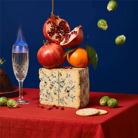 A cheese and fruit still life with pomegranate, orange, and Brussels sprouts on a red tablecloth and colorful background.