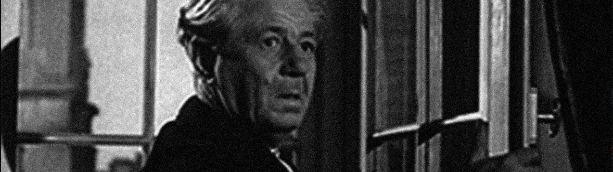 A black and white photo of an elderly woman with short, styled hair looking through a window with another person shadowed inside.