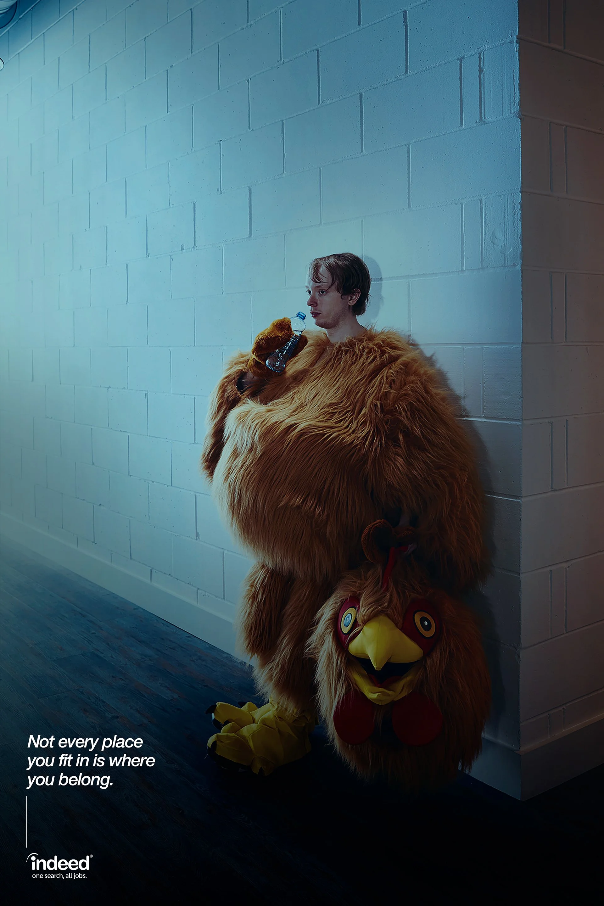 A young man wearing a lion costume with a large lion head mask on the ground next to him, holding a water bottle, standing against a white brick wall in a dimly lit area.