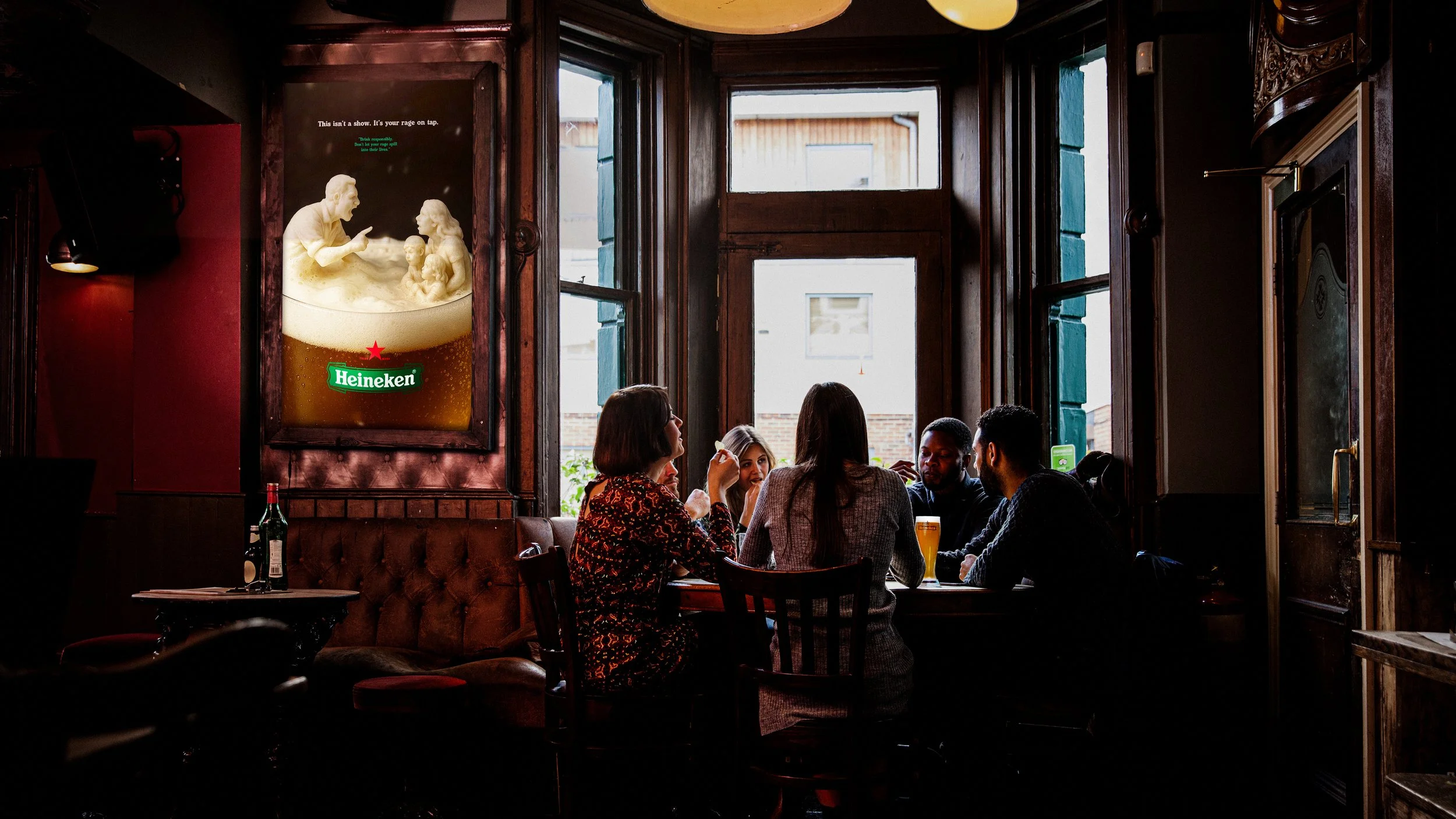 People sitting at a table in a dimly lit pub or restaurant with wooden decor. Four women and two men are engaged in conversation, with drinks on the table. A large Heineken beer advertisement featuring a foamy beer with a family is visible on the wall. Sunlight is streaming in through tall open windows.