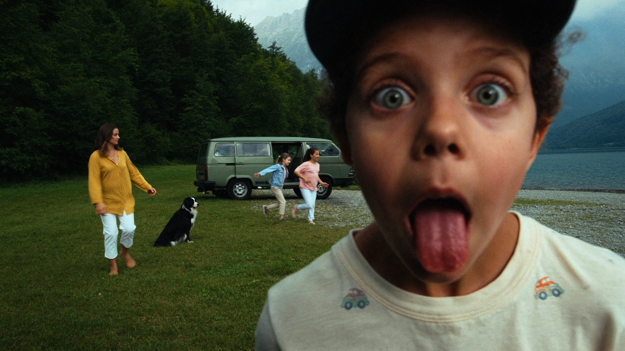 Close-up of a young boy with wide eyes and tongue sticking out in front of a scenic outdoor setting with a lake, green trees, and mountains. In the background, a woman, two girls, and a dog are visible near a green van.