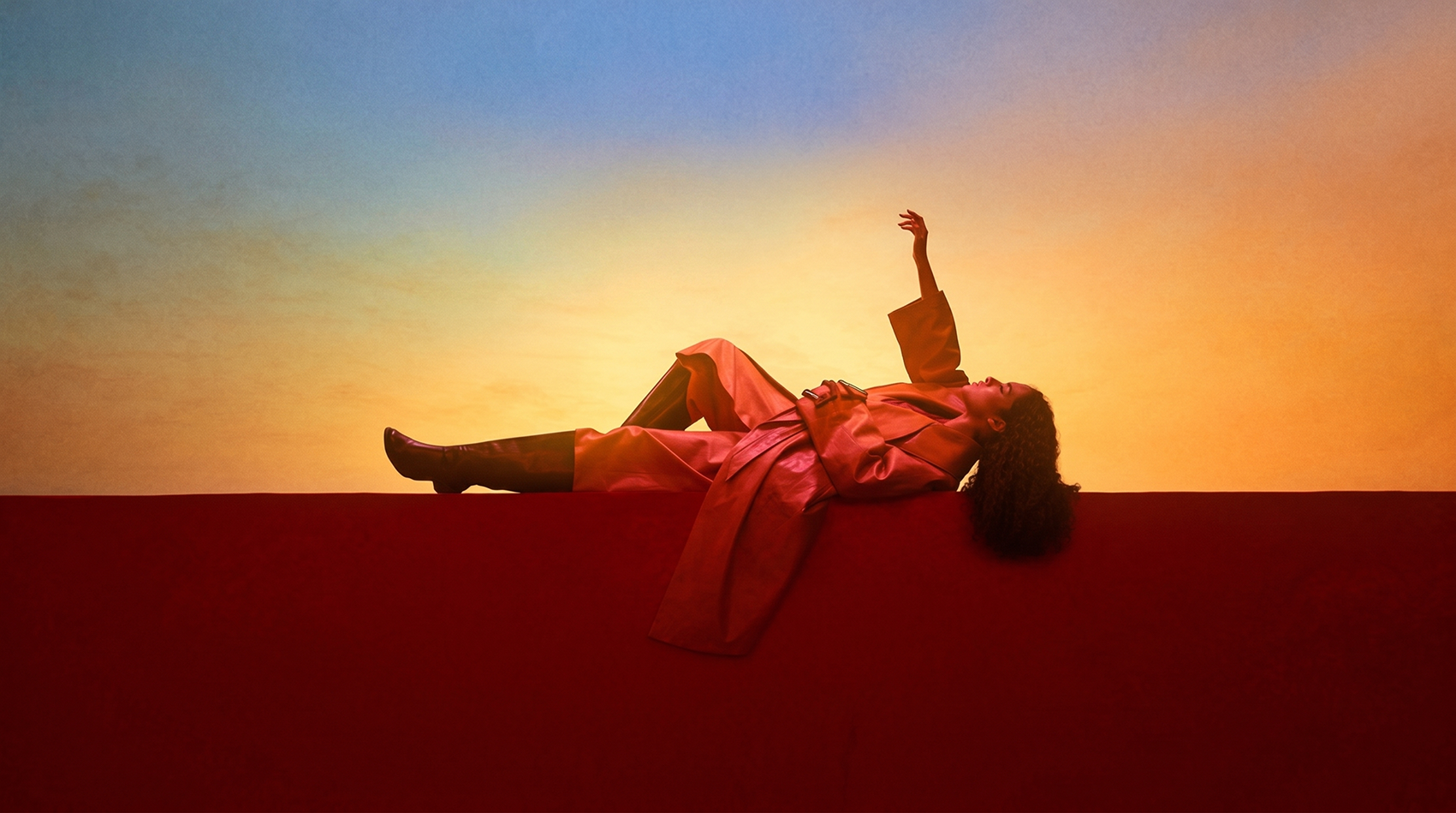 A woman with curly hair lying on a red surface with a sunset-like gradient sky in the background, wearing a trench coat, with one arm raised and gazing upward.