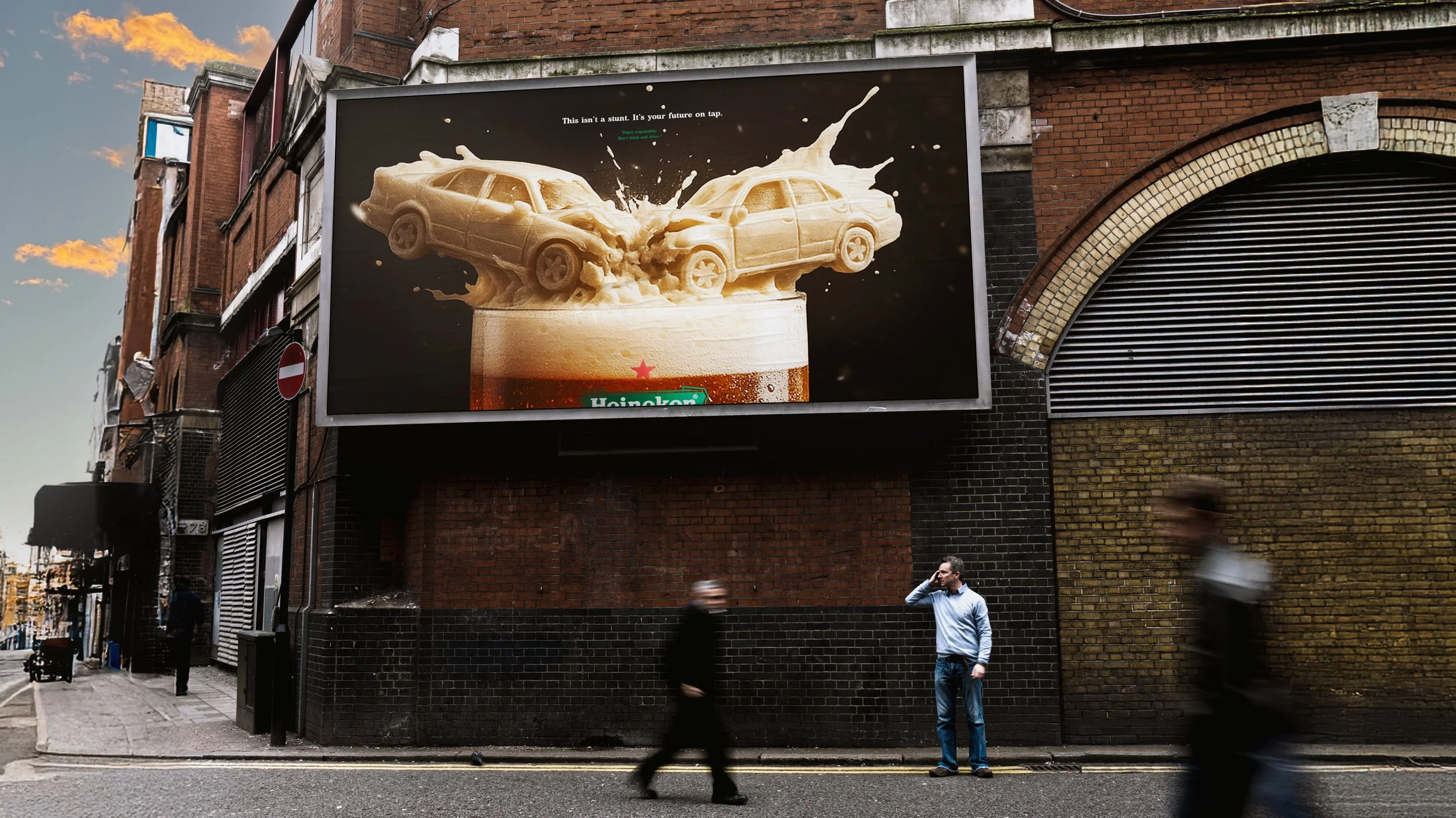 Billboard on a city street showing a large image of two cars made of beer foam crashing into each other, with a Heineken beer glass underneath. The billboard reads: 'This isn’t a stunt. It’s your future on tap.' Pedestrians walking past at sunset.