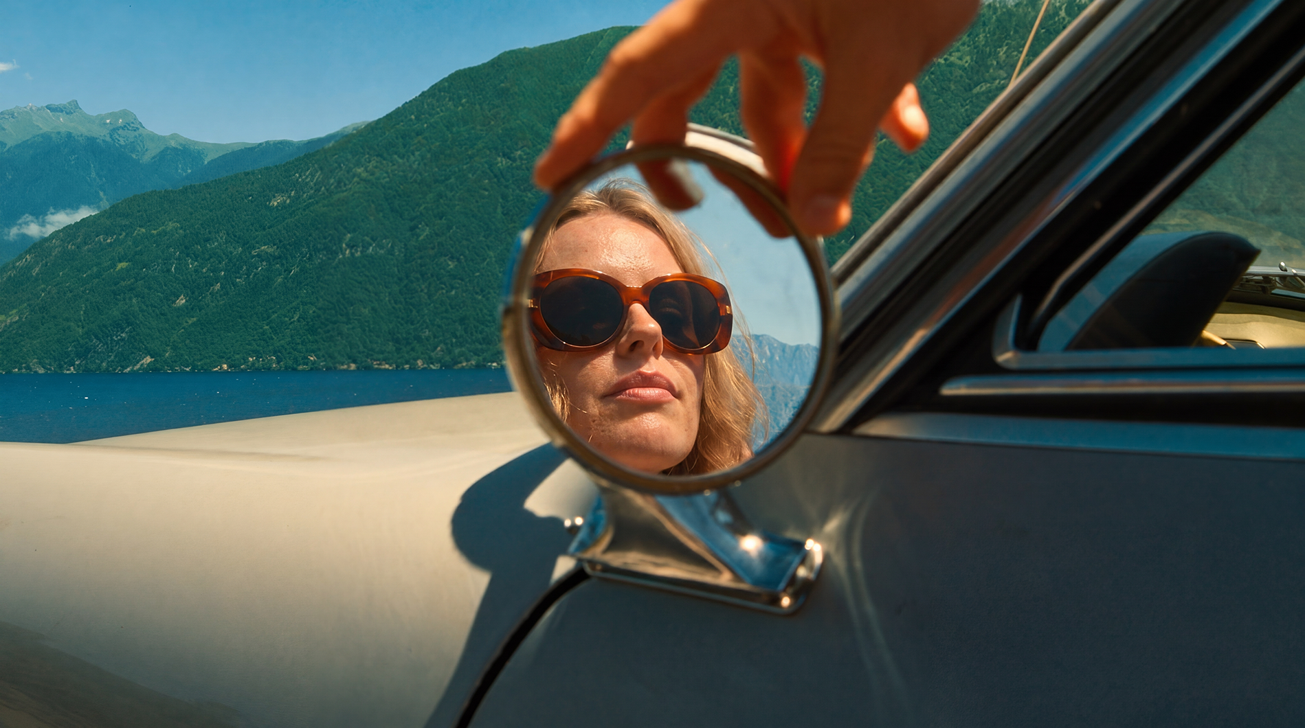 Woman with sunglasses looking into a small mirror held by hand outside a car, with mountains, lake, and blue sky in the background.