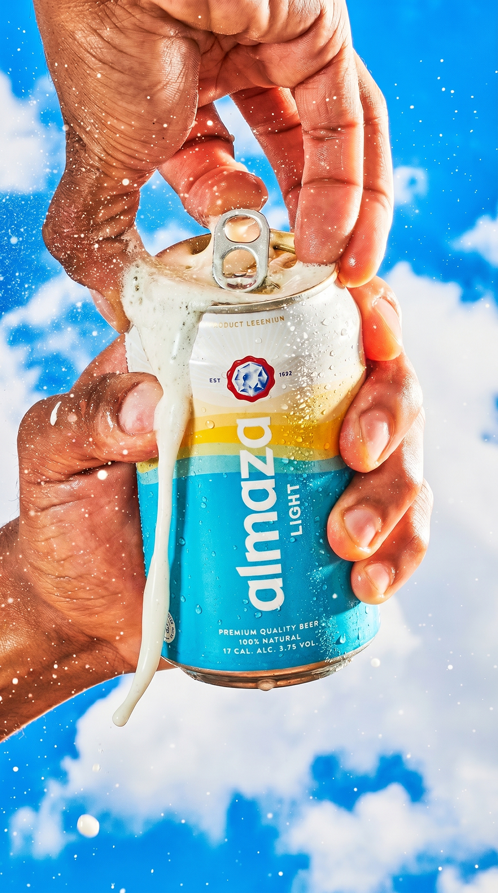 Person opening a can of Malibu Light beer with foam overflowing, against a backdrop of blue sky and clouds, with snow or confetti in the air.