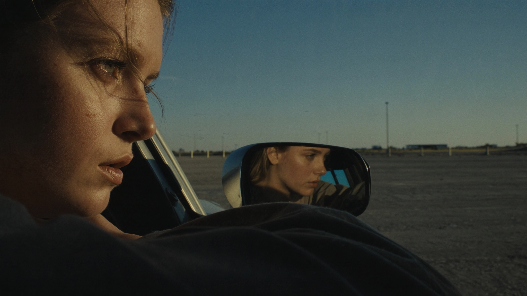 A woman in a car with a young woman in the side mirror, both looking serious, on an empty road during sunset.