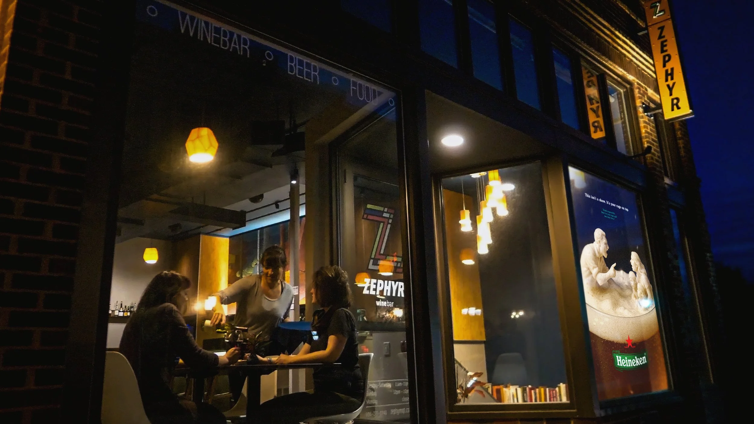 People dining inside Zephyre wine bar at night, visible through a large glass window with a neon Heineken sign outside, some patrons are ordering or talking, warm lighting inside, urban street setting.