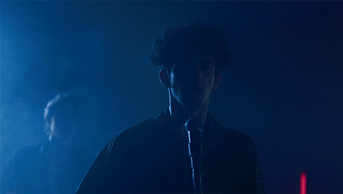 A person with curly hair singing into a microphone on stage with blue lighting, with another person visible in the background.
