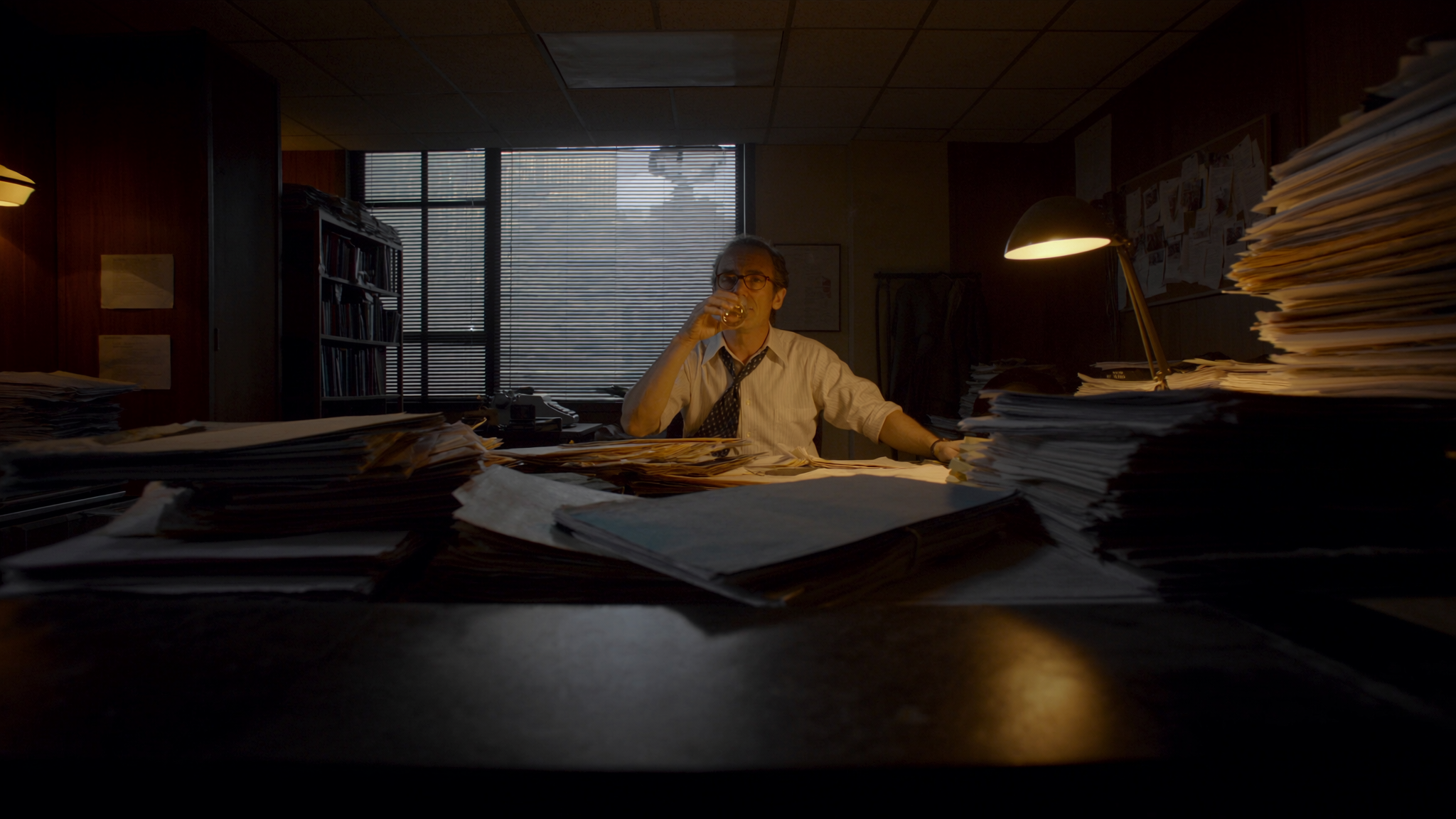 Man sitting at cluttered desk drinking from a glass, office with large window, wooden walls, shelves with books, and a lamp casting yellow light.