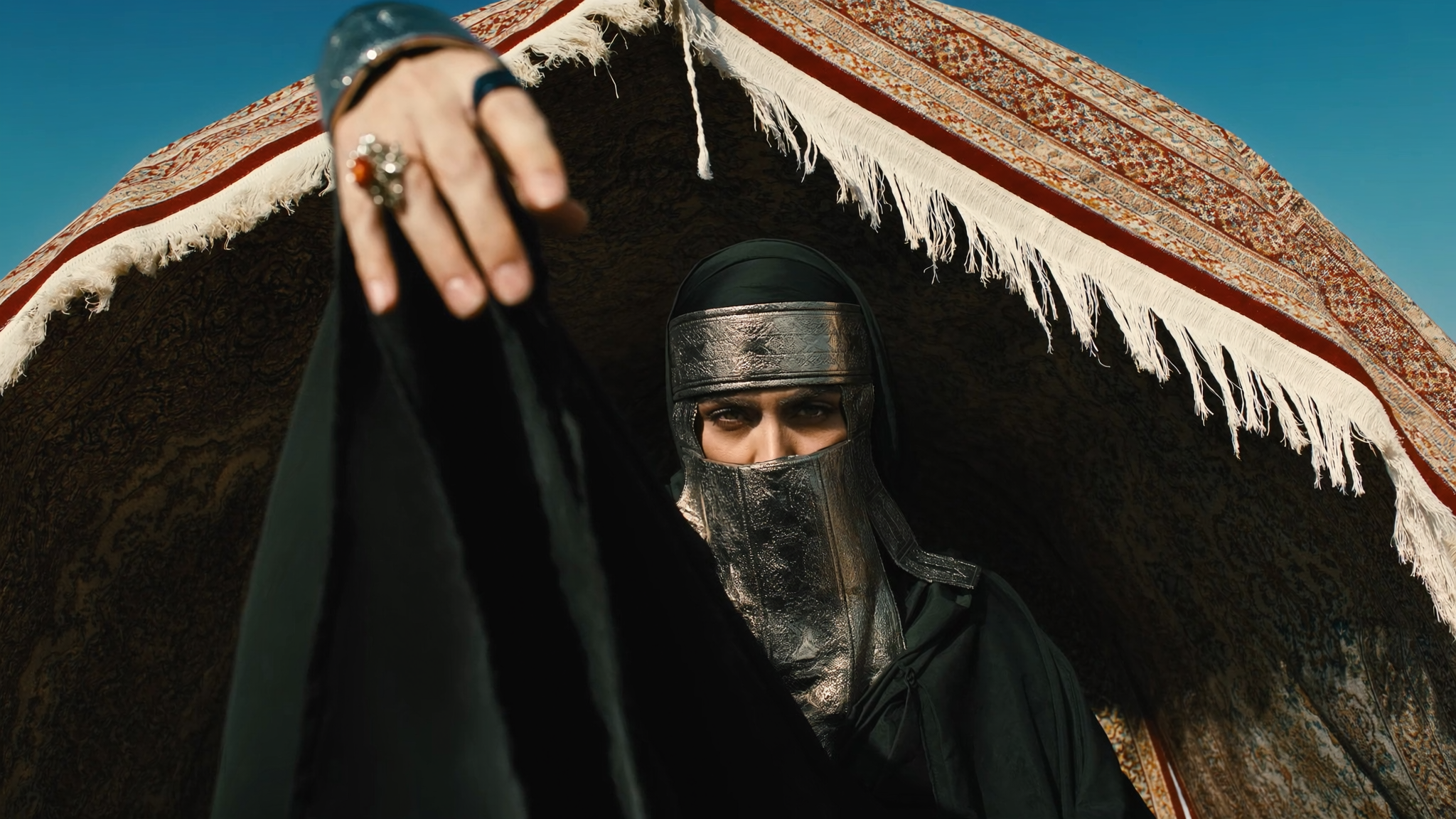 A person dressed in medieval armor and clothing, wearing a metallic helmet and a cloth mask, is partially visible underneath a large ornate carpet or rug held above their head with fringes hanging down, against a clear blue sky background.