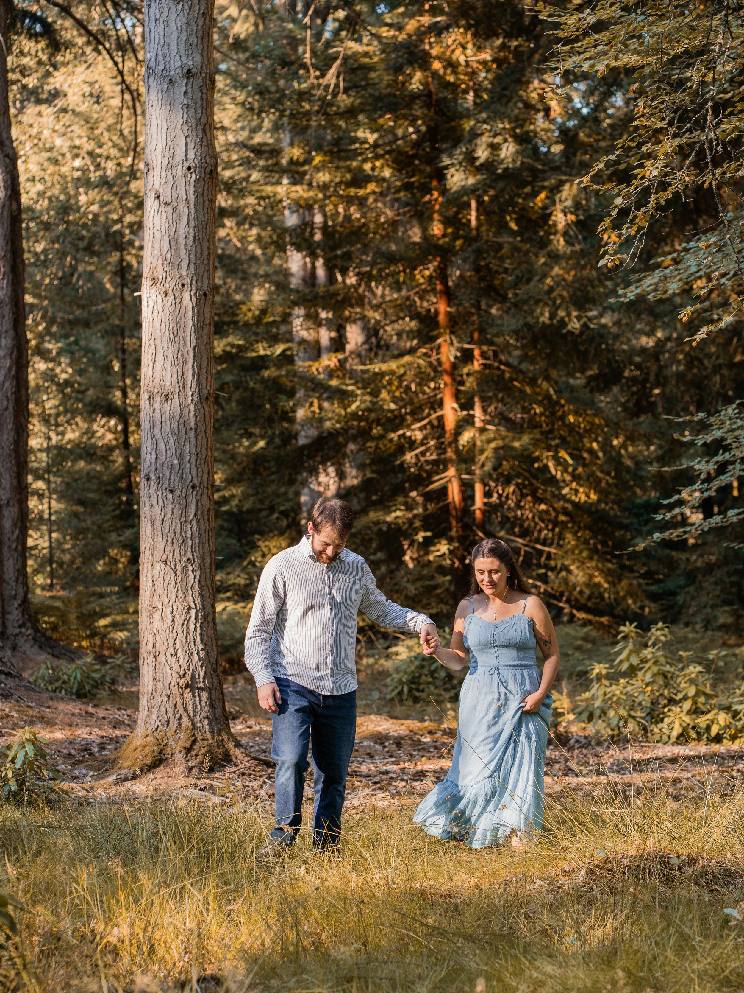 A couple is walking through a forest during sunset, holding hands and smiling.