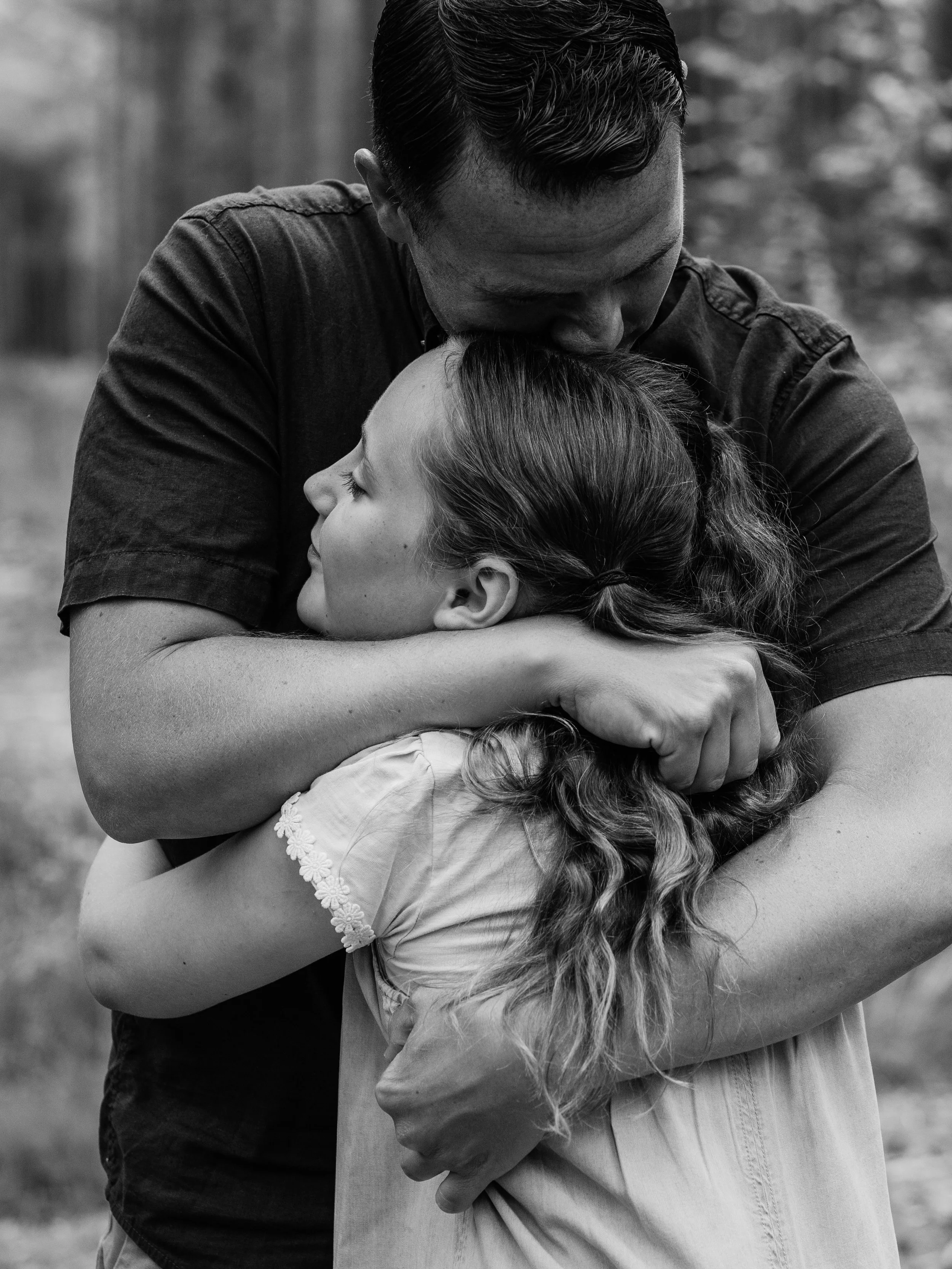 A black and white photo of a man embracing a young girl tightly, with his face close to her head and eyes closed. The girl has long hair and is smiling softly, with her eyes closed, leaning into the embrace.