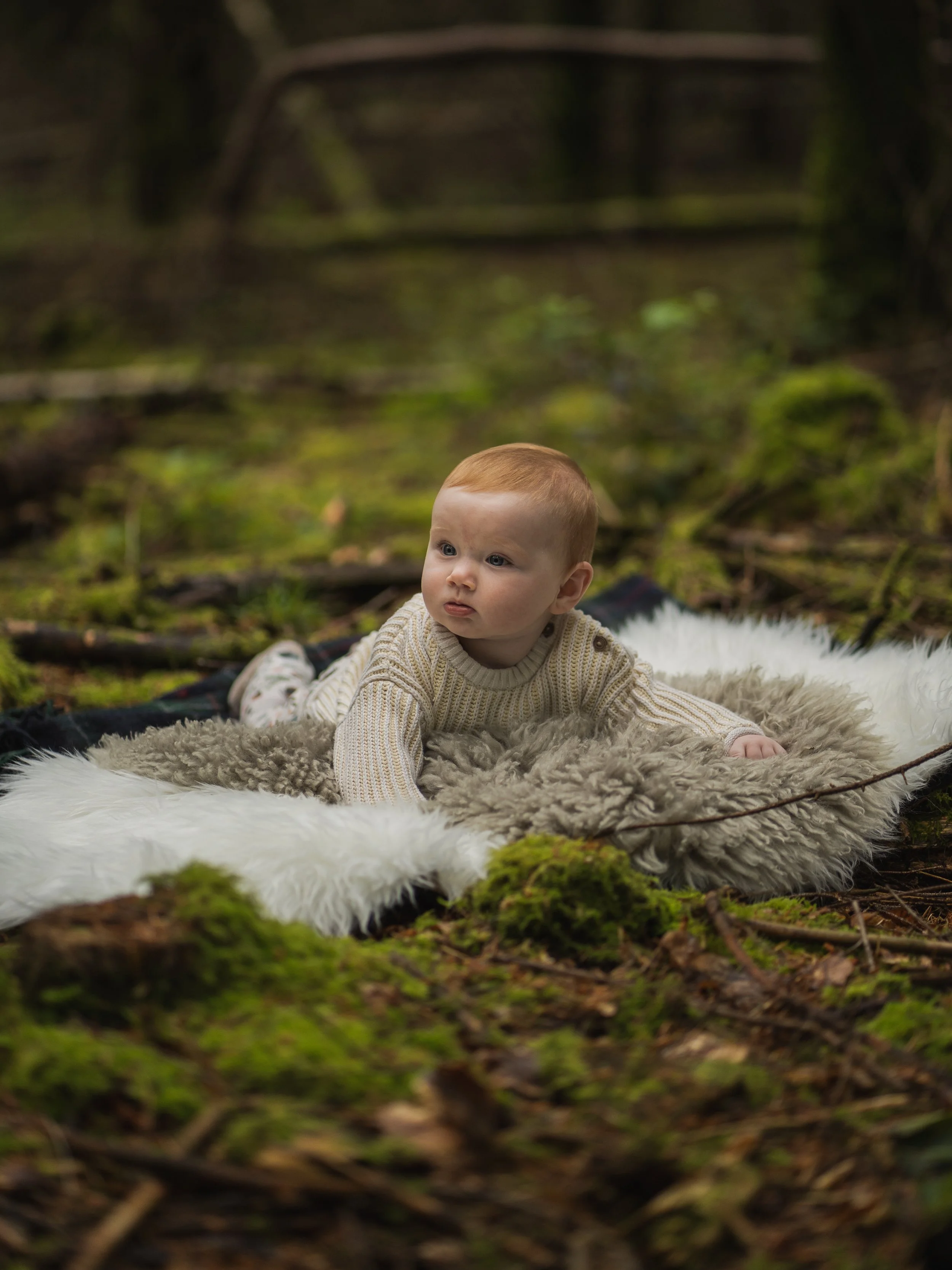 A young child with red hair and blue eyes, wearing a beige knitted sweater, lying on a white furry blanket in a forest setting with moss-covered ground and blurred trees in the background.
