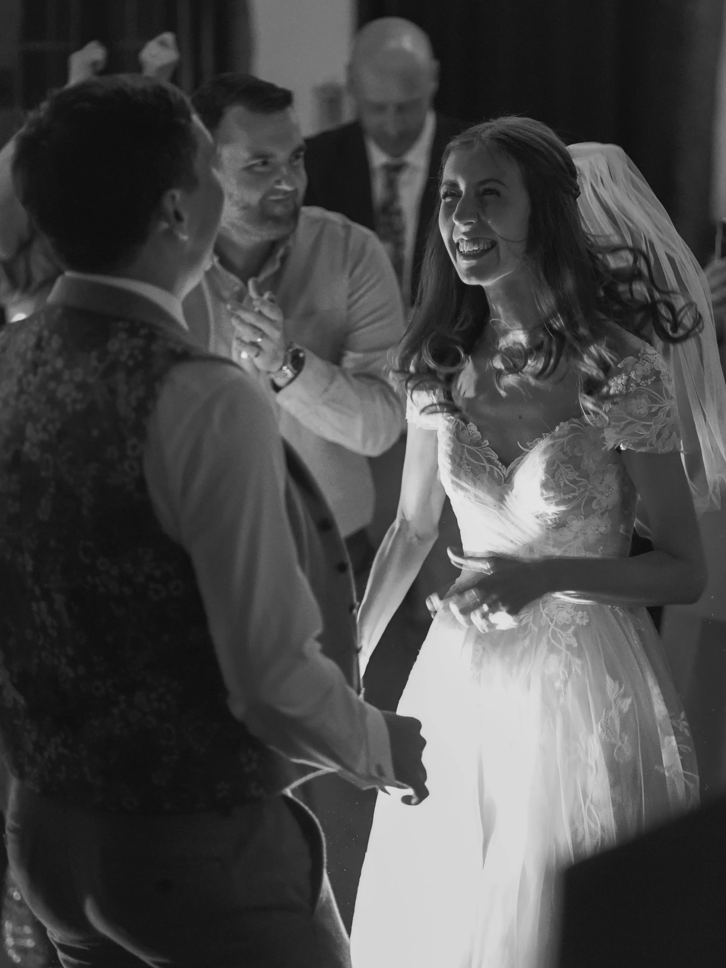 Black and white photo of a wedding scene with a bride in a white dress and veil, smiling and holding hands with a groom, during their wedding dance. Three other men are in the background, one with a beard and glasses smiling, and two others facing ea