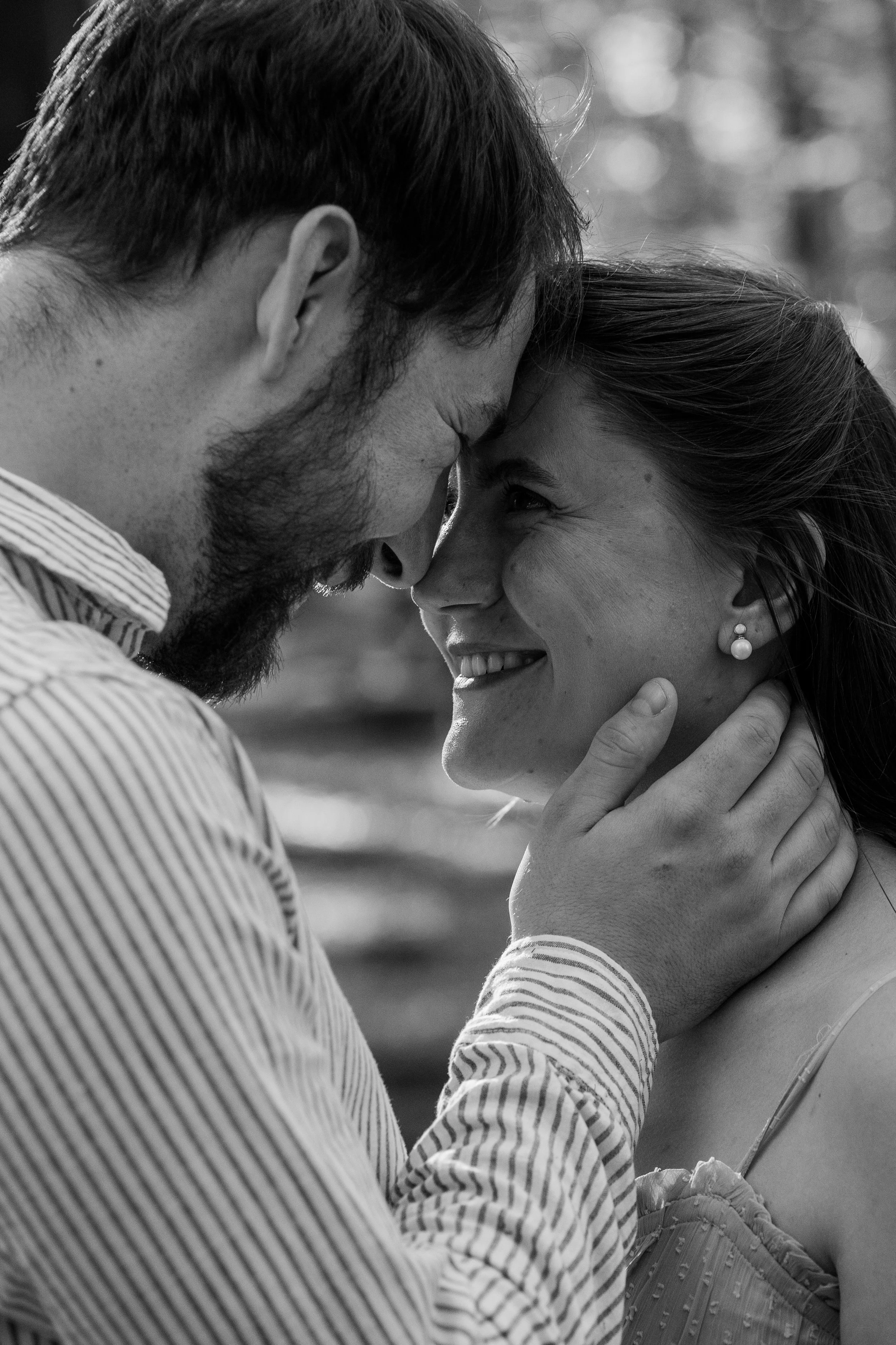 A black-and-white close-up photograph of a couple touching foreheads and smiling. The man has dark hair, a beard, and is wearing a striped shirt. The woman has dark hair, earrings, and is wearing a blouse with a textured pattern.