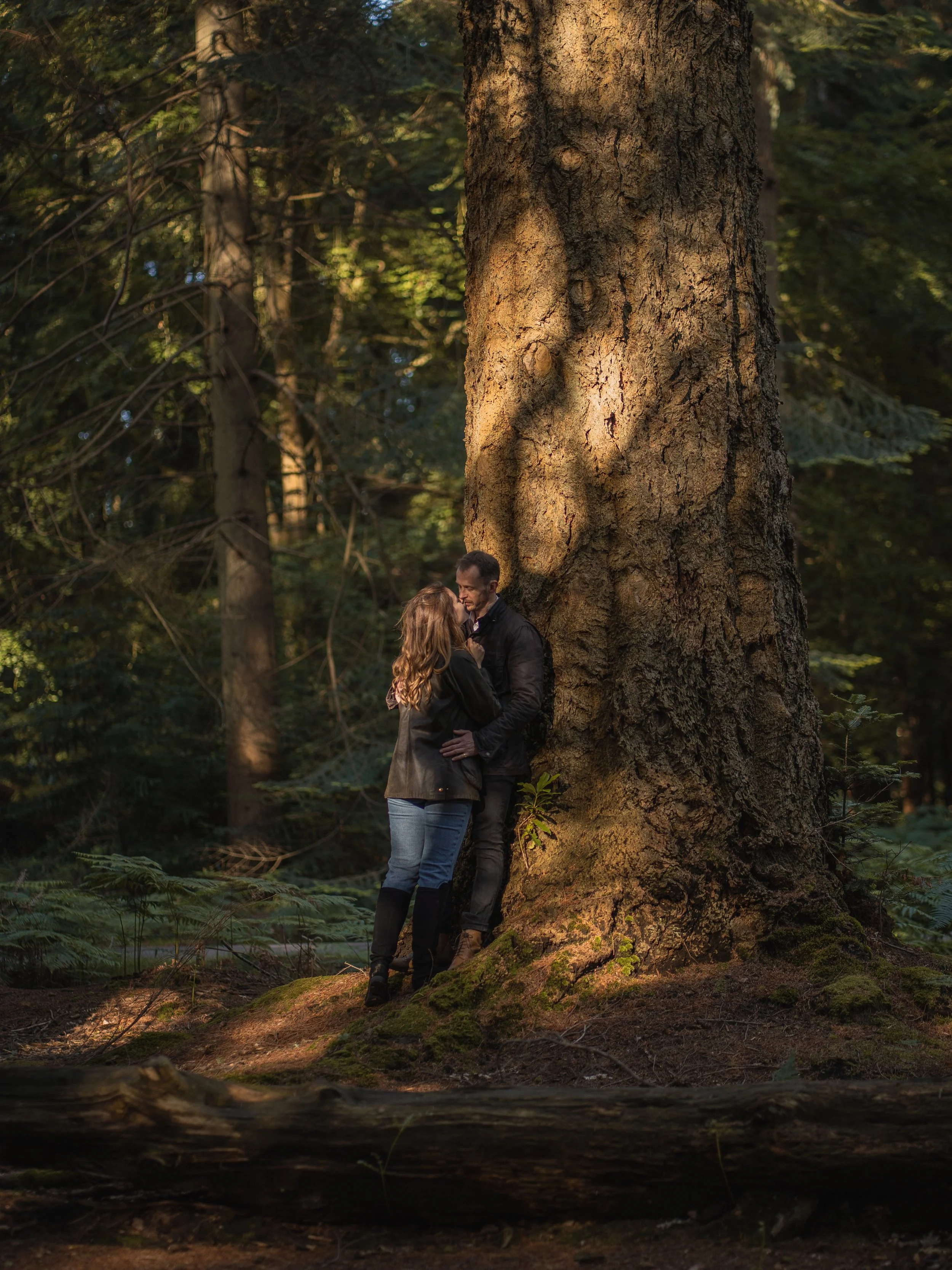 A man and woman stand close together in a forest, near a large tree, with sunlight filtering through the trees in the background.