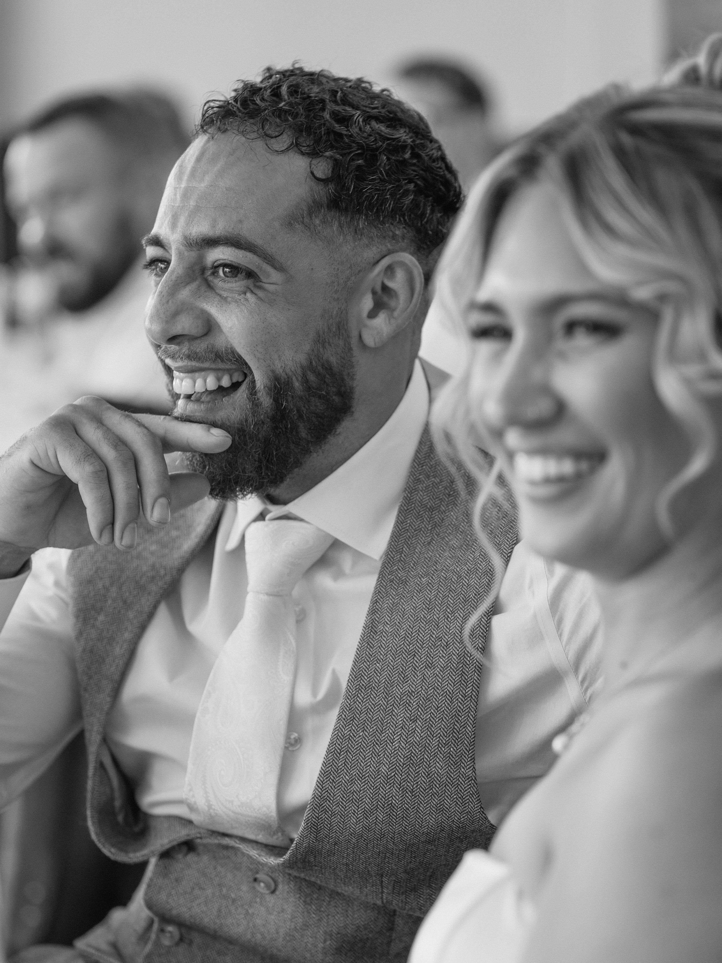 Black and white photo of a man in a suit and tie smiling and looking to the side, with people blurred in the background, attending a formal event.