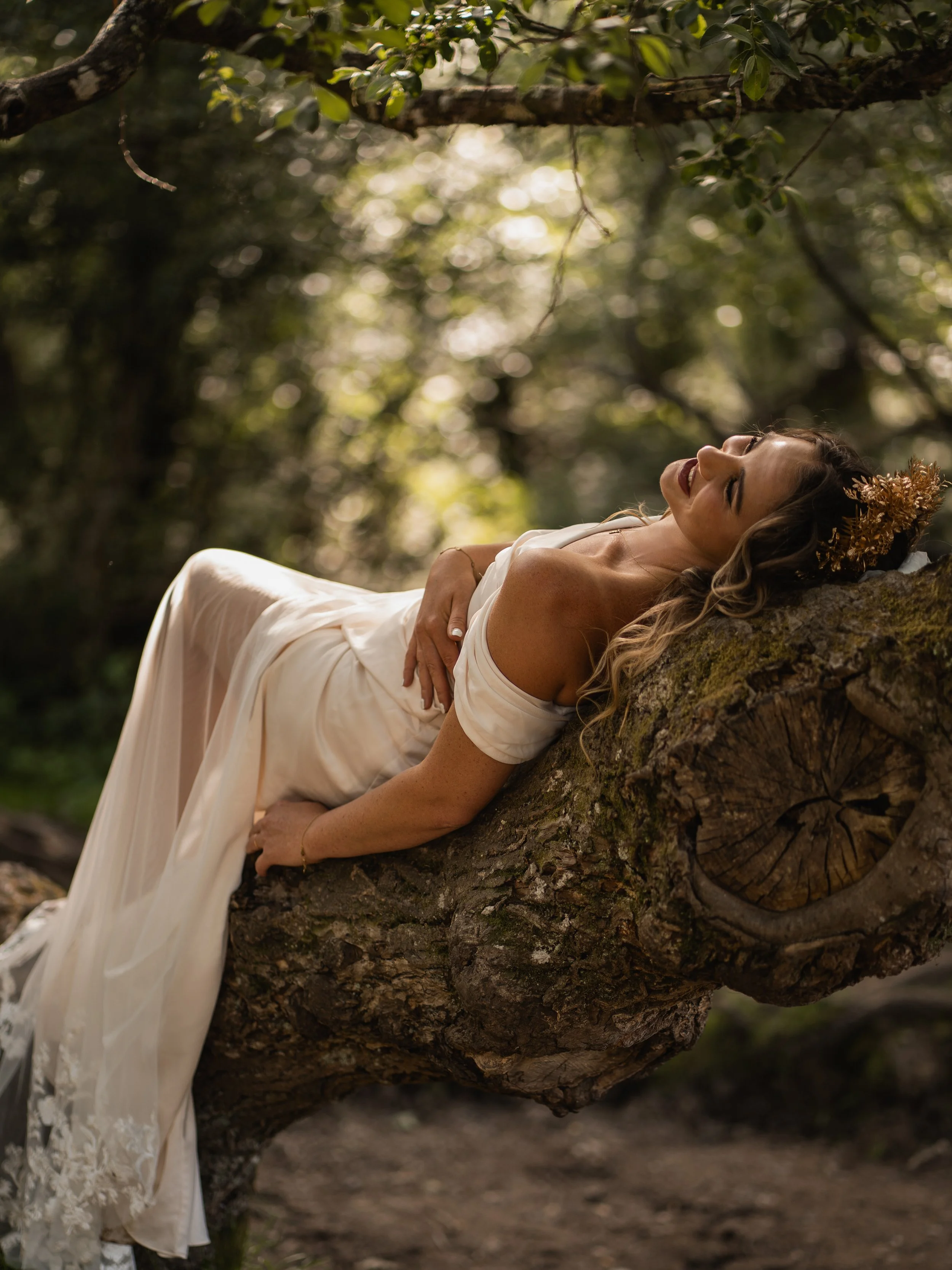 Woman in a white dress lying on a large tree branch in a wooded area, smiling and looking up at the sky.