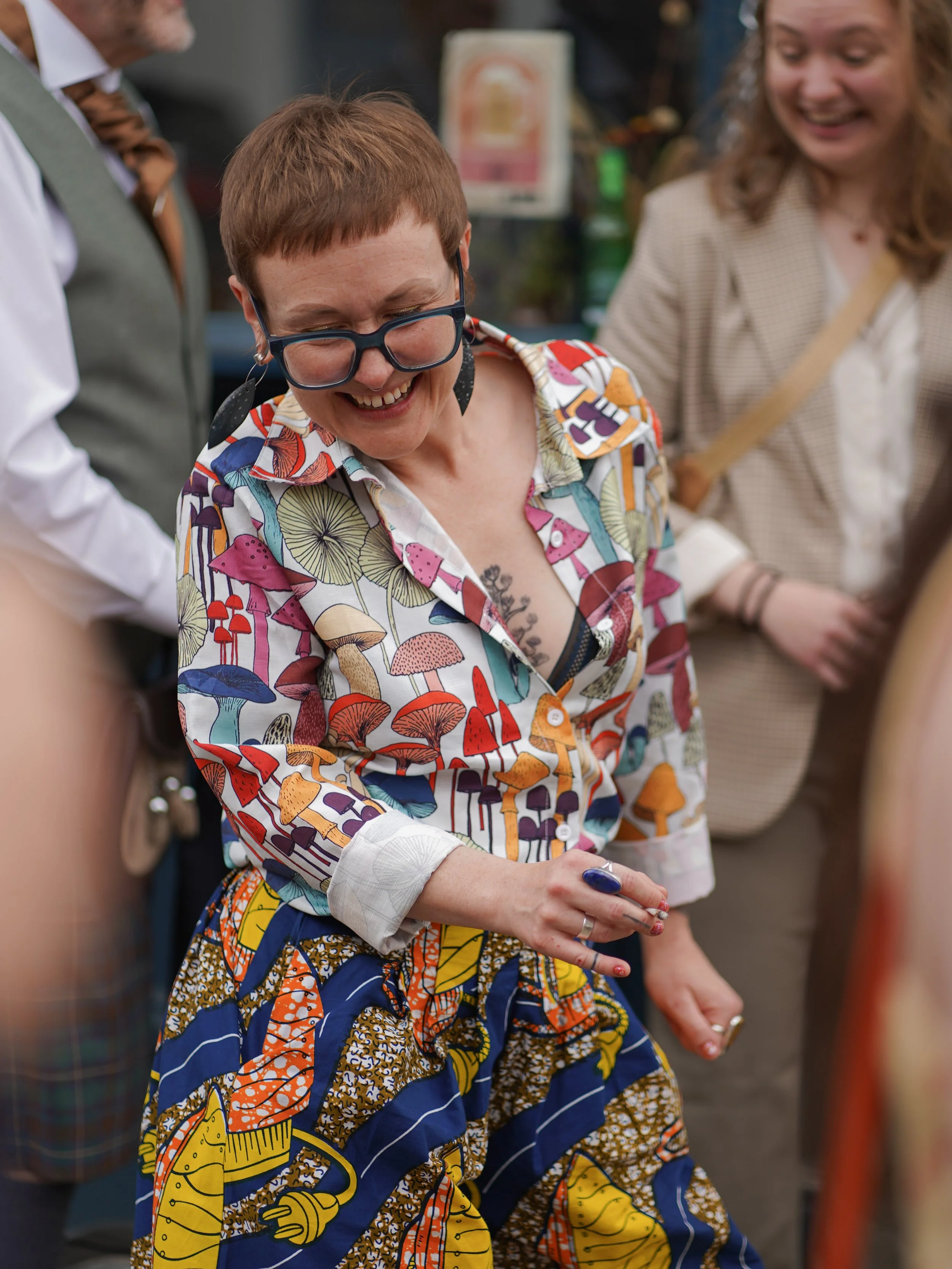A woman with short red hair, wearing glasses, in a colorful embroidered shirt, smiling and looking down, holding a pen. Two other women are partially visible beside her, one in a beige checked blazer and another with a white shirt and tie, possibly o