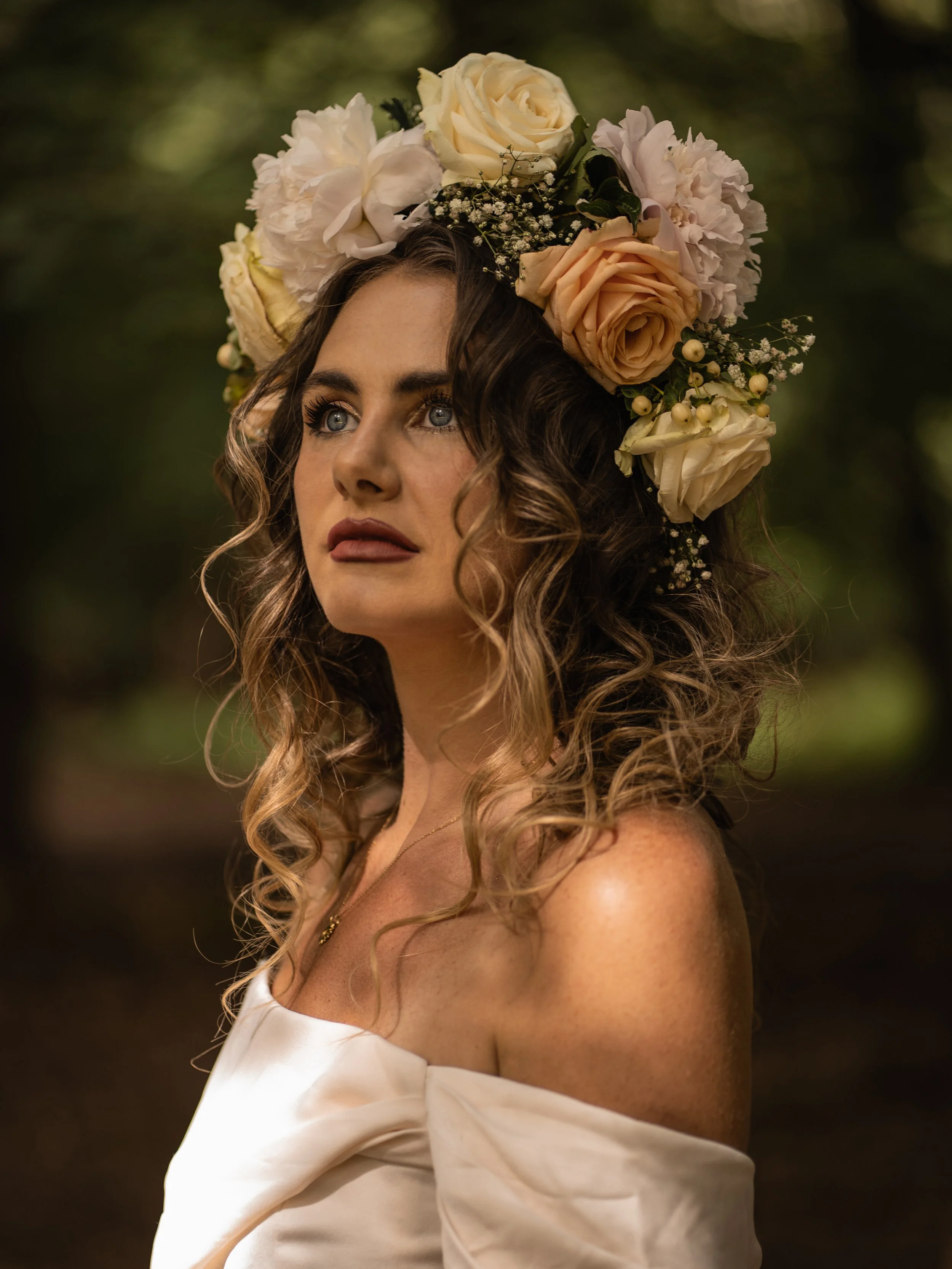 A woman with curly brown hair wearing a large floral crown made of white and peach roses, white peonies, baby's breath, and greenery, outdoors with a blurred green background.
