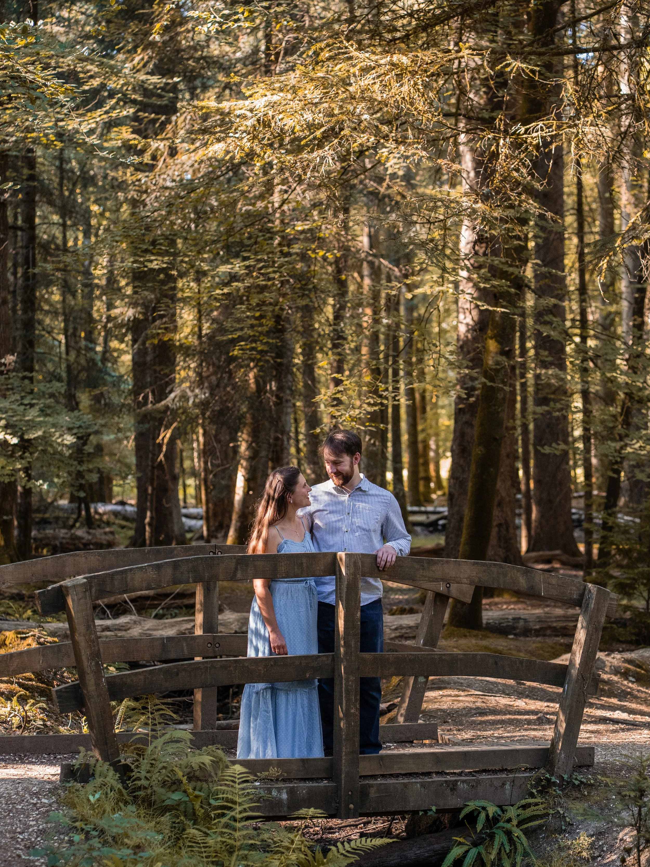 A couple stands on a wooden bridge in a forest, looking at each other and smiling, surrounded by tall trees and green foliage.