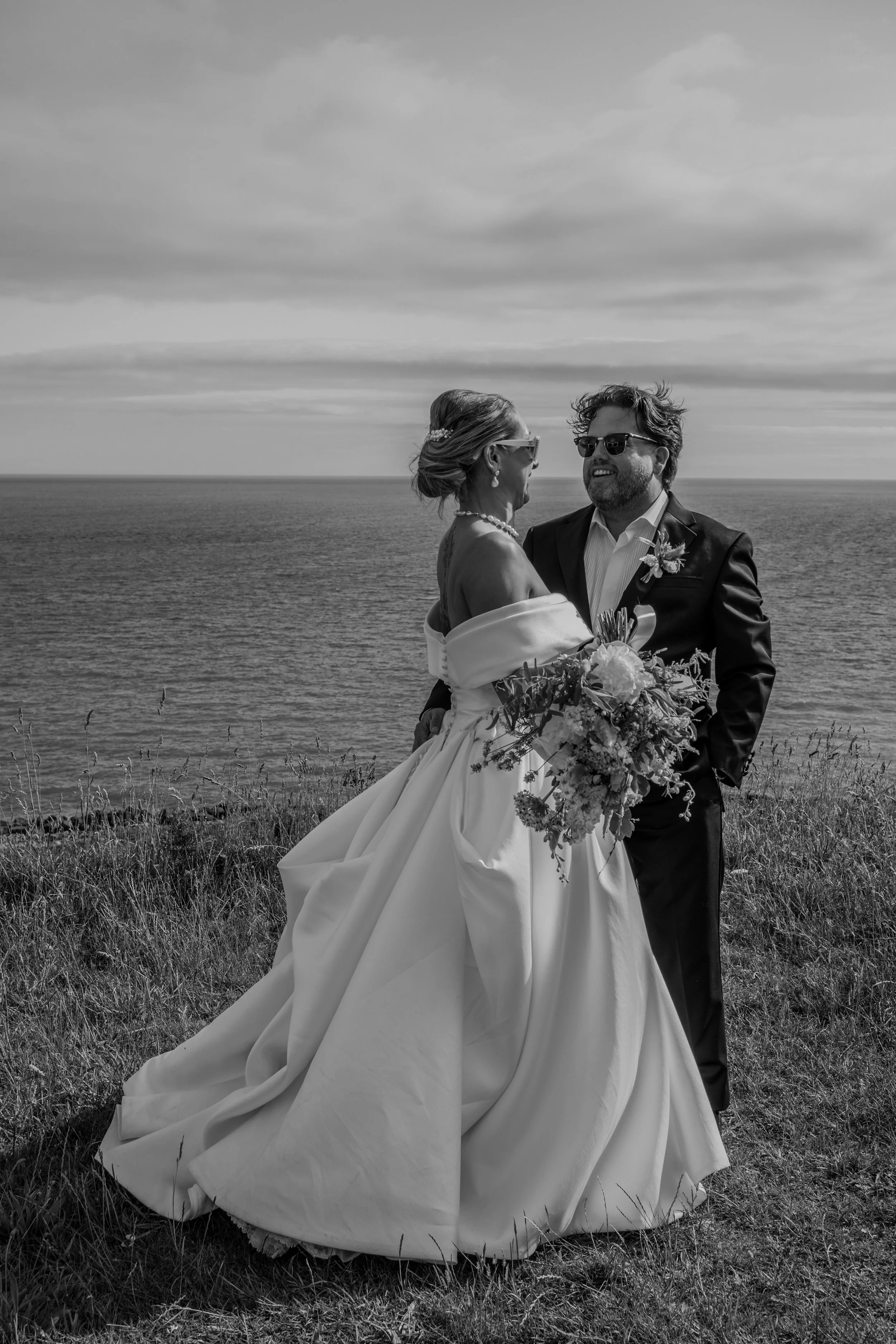 A black and white photo of a wedding couple standing outdoors near the ocean, with the bride in a wedding gown and the groom in a suit, sharing a joyful moment.