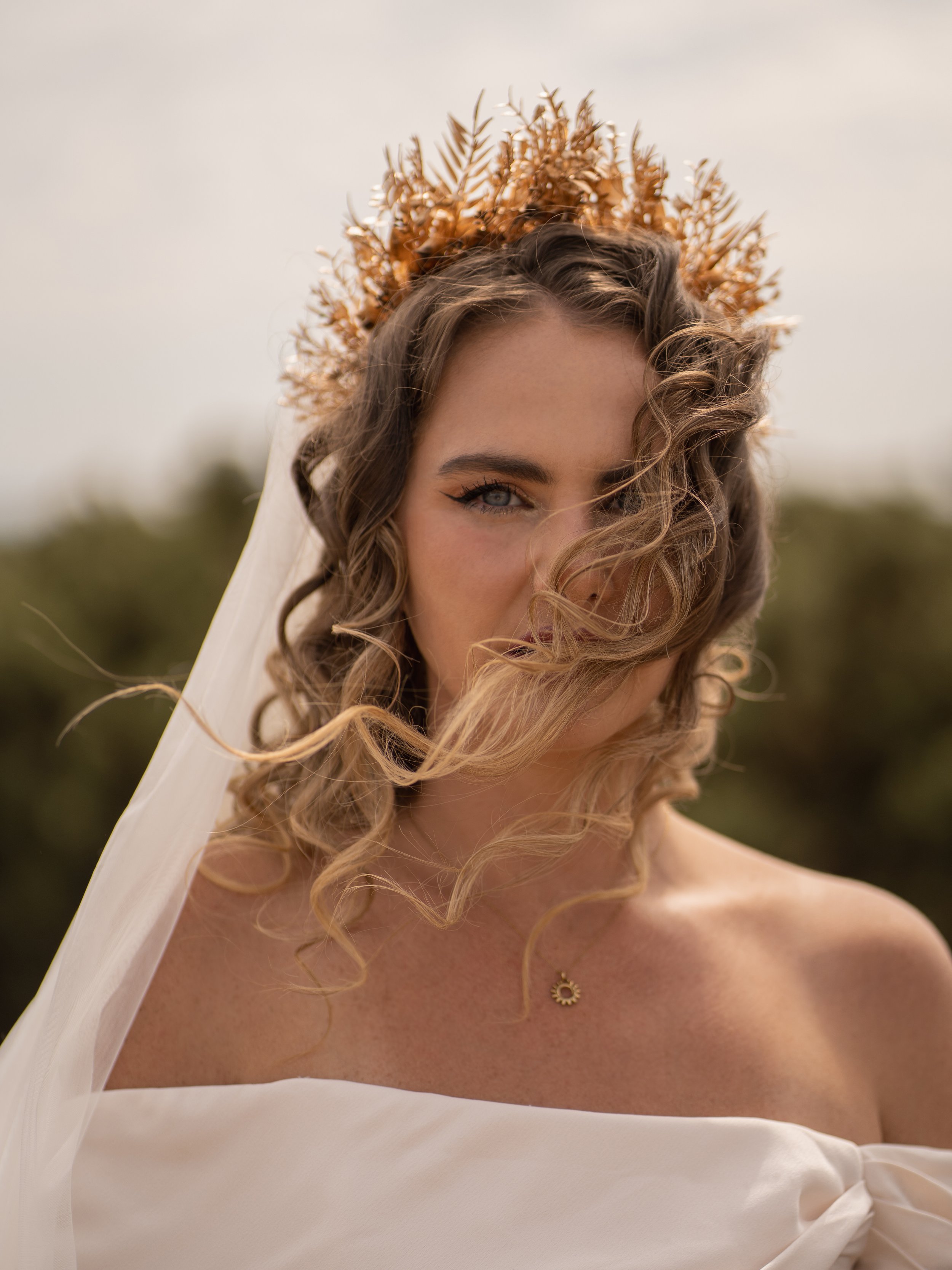 Close-up of a woman with curly hair wearing a cream-colored off-the-shoulder dress, a gold necklace, and a crown made of dried leaves, outdoors during daytime.