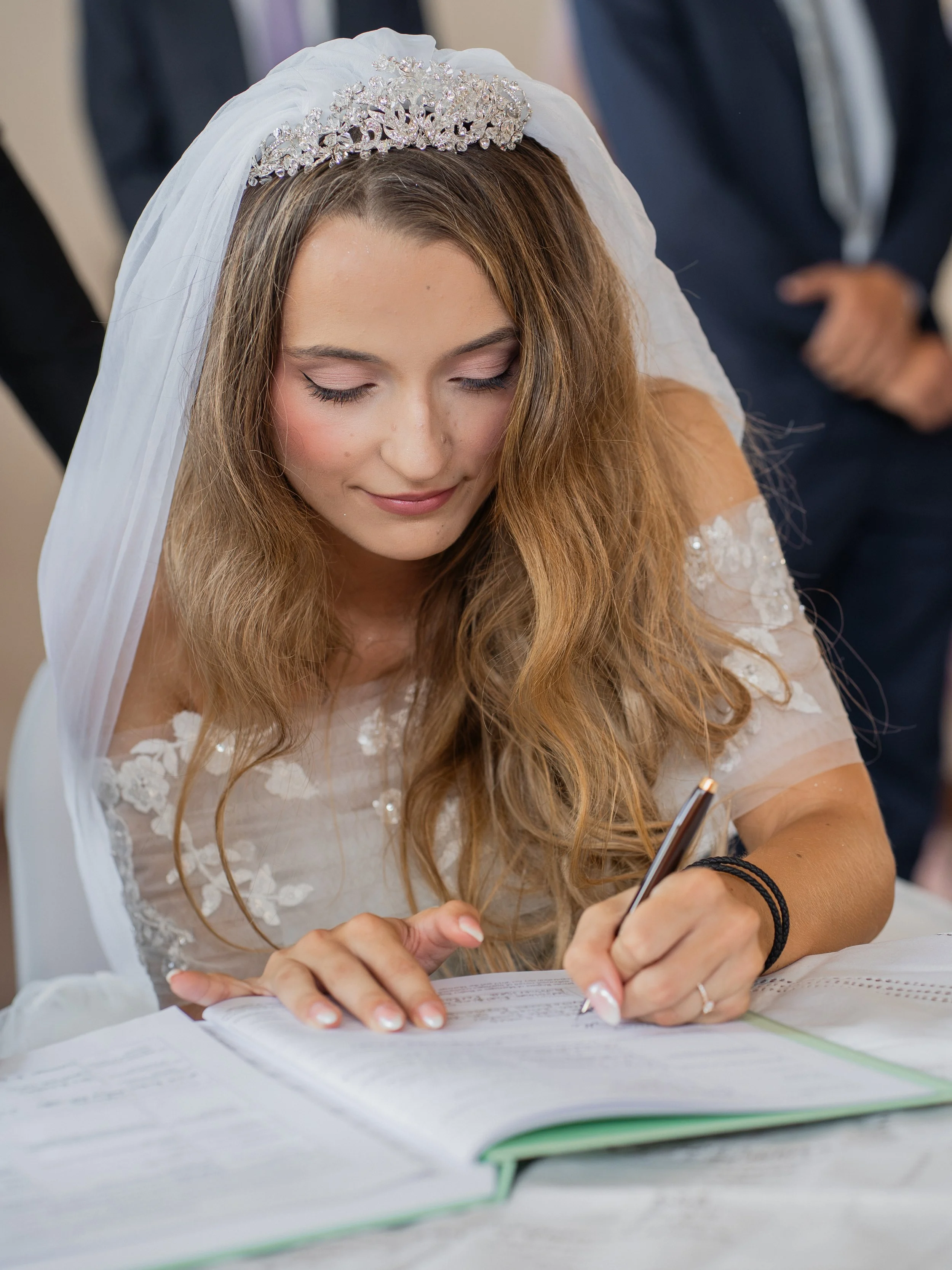A woman with wavy hair and a tiara and white bridal veil is smiling and signing a document with a pen. She is wearing a white lace dress with floral embroidery and has black bracelets on her wrist.