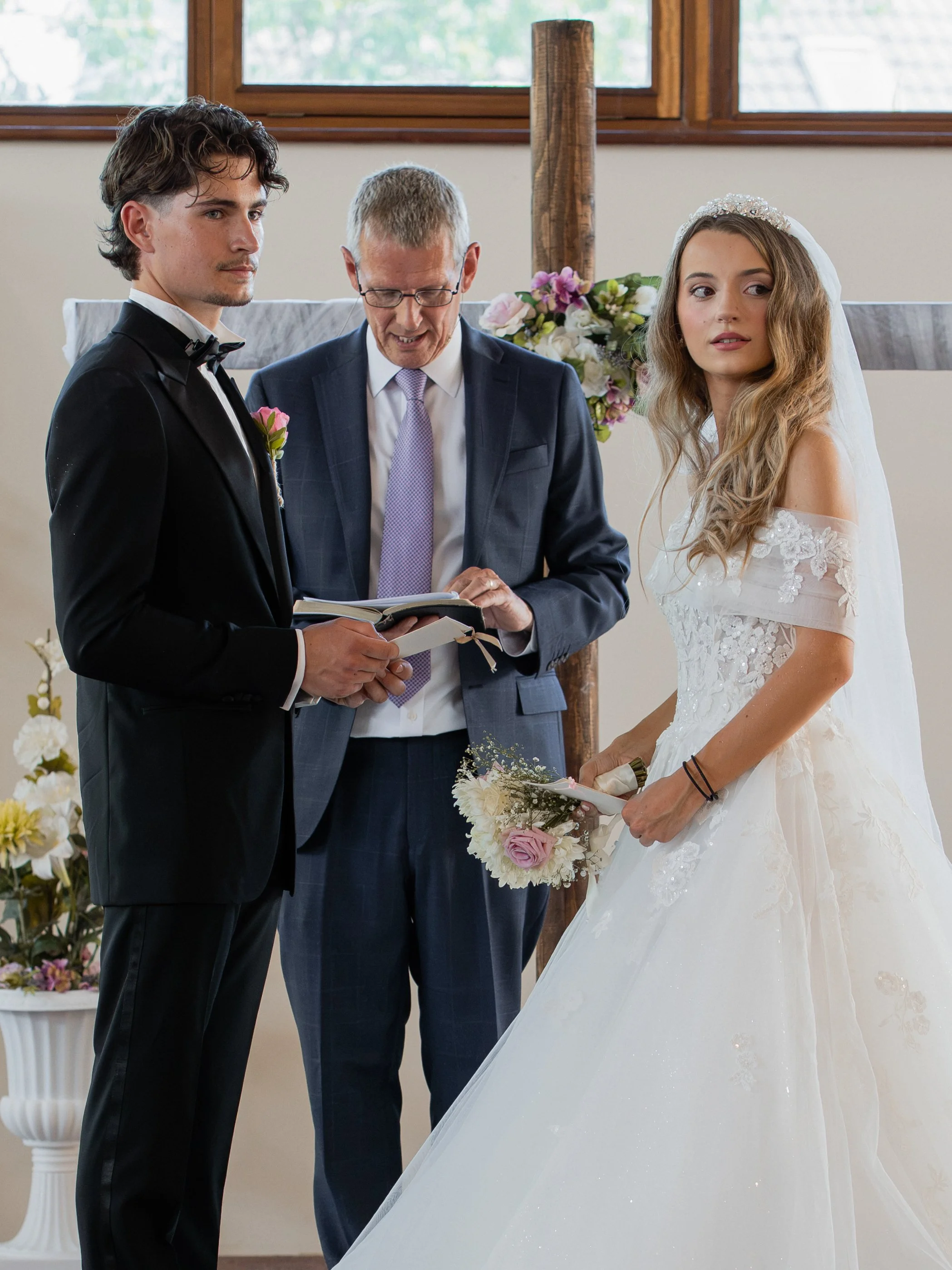 A wedding ceremony with a bride and groom standing opposite each other, facing a officiant. The bride is holding a bouquet of flowers, and the groom is dressed in a black tuxedo. The officiant is reading from a book. The setting features large window