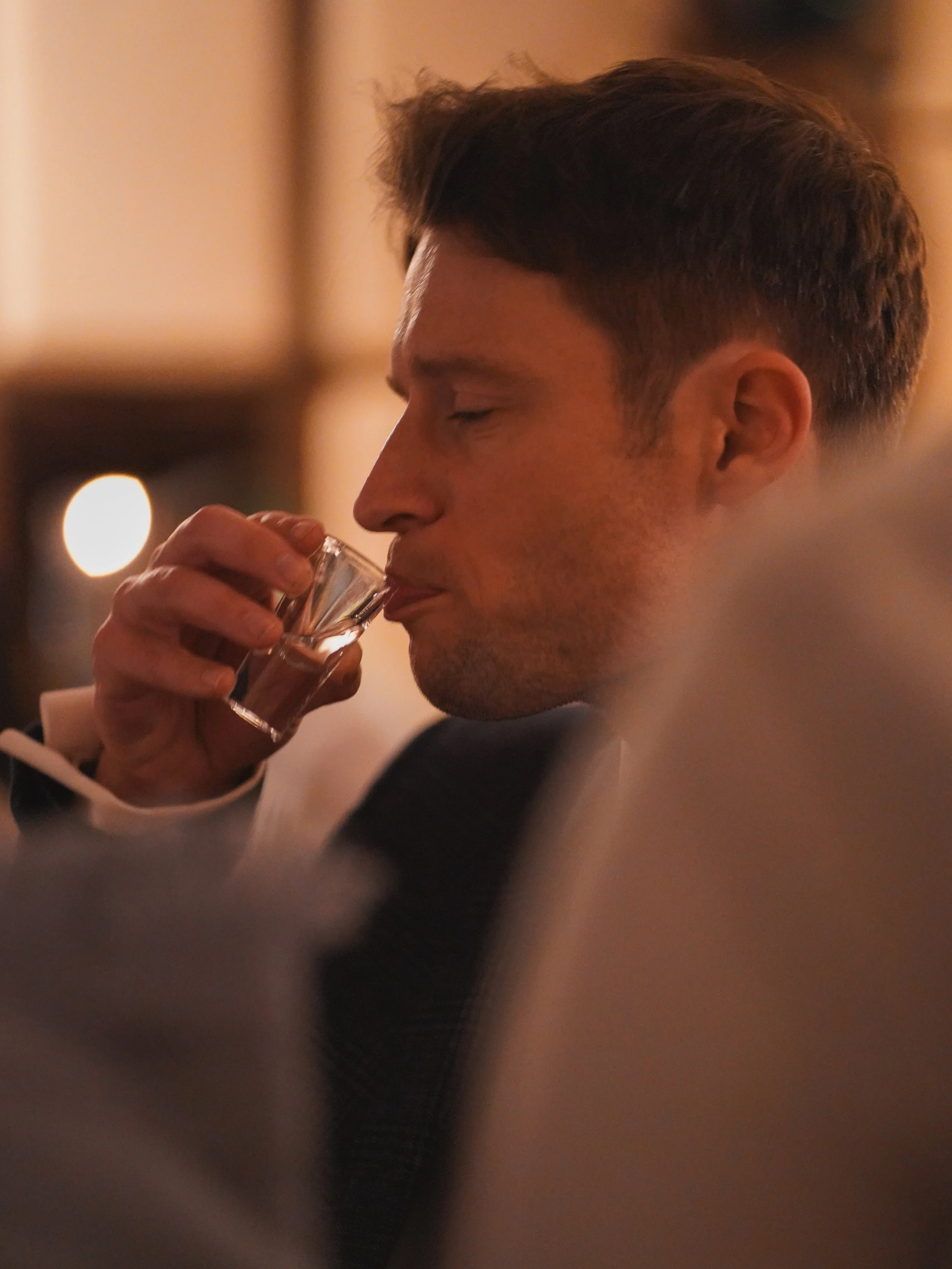 A man with short dark hair drinking from a shot glass in a dimly lit setting.