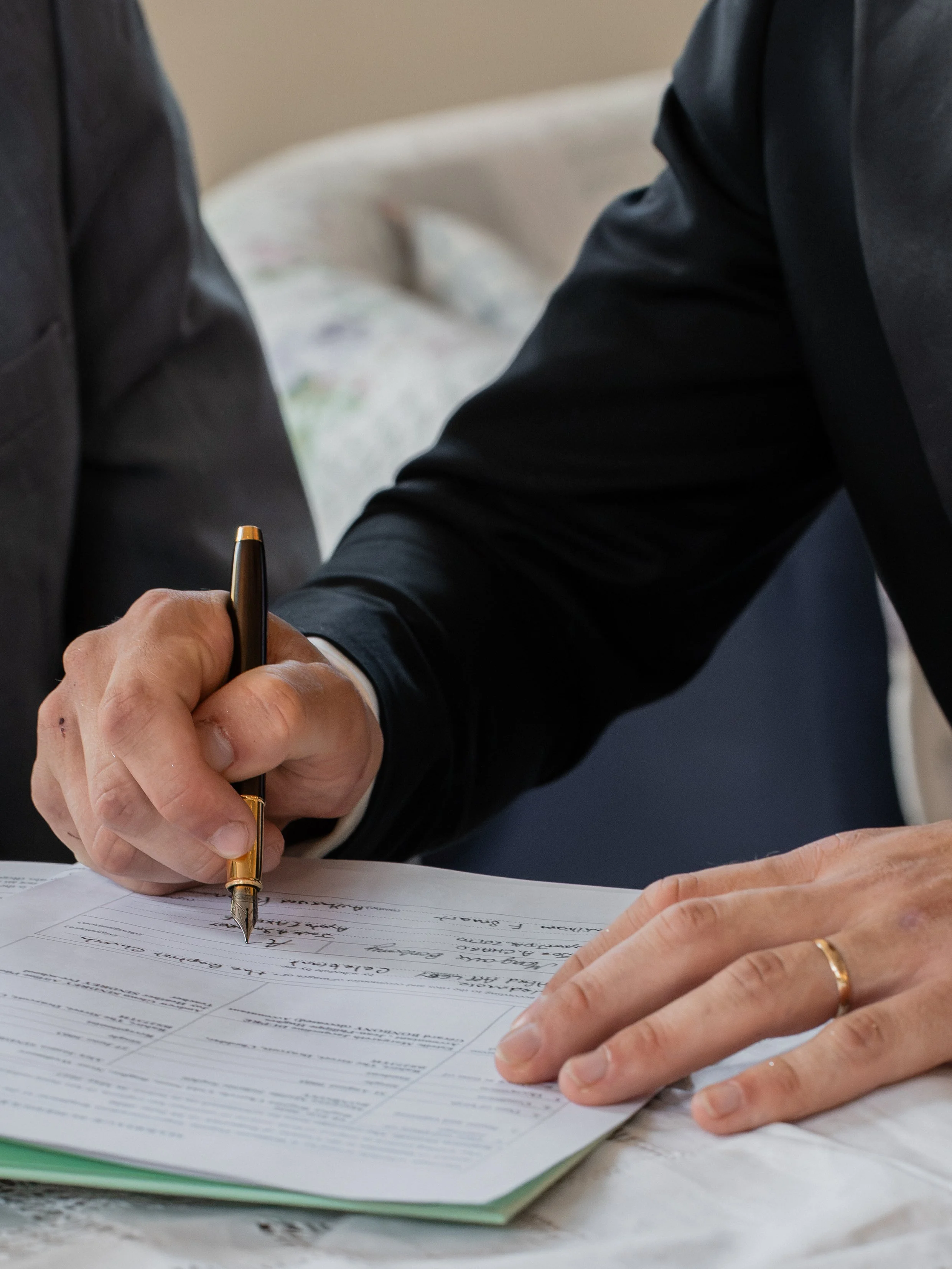 Close-up of two people signing a document with a gold and black pen. One person is wearing a dark suit and a gold ring, the other person has a gold ring on their finger.