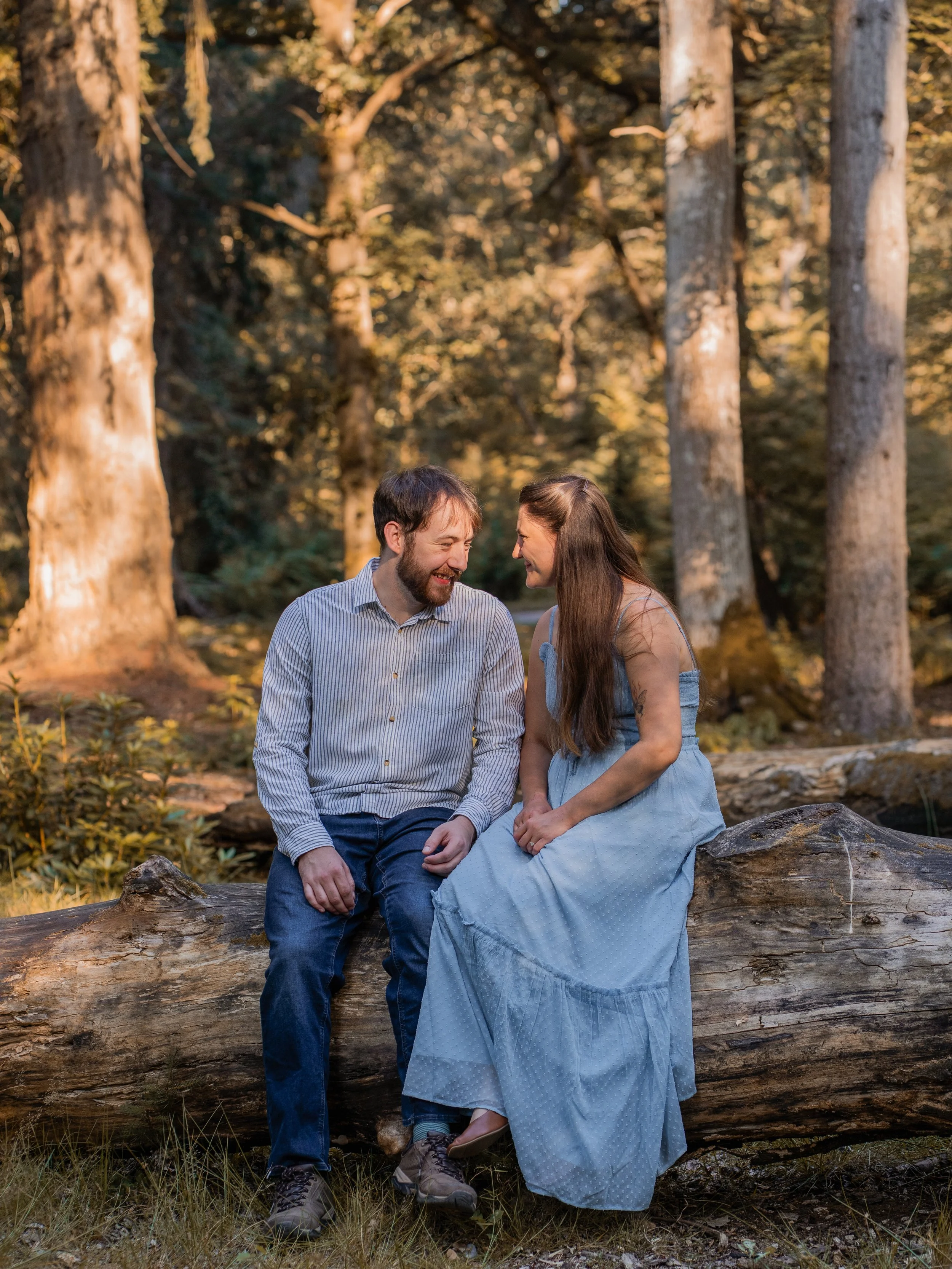 A man and woman sitting on a fallen log in a forest during sunset, smiling and looking at each other.