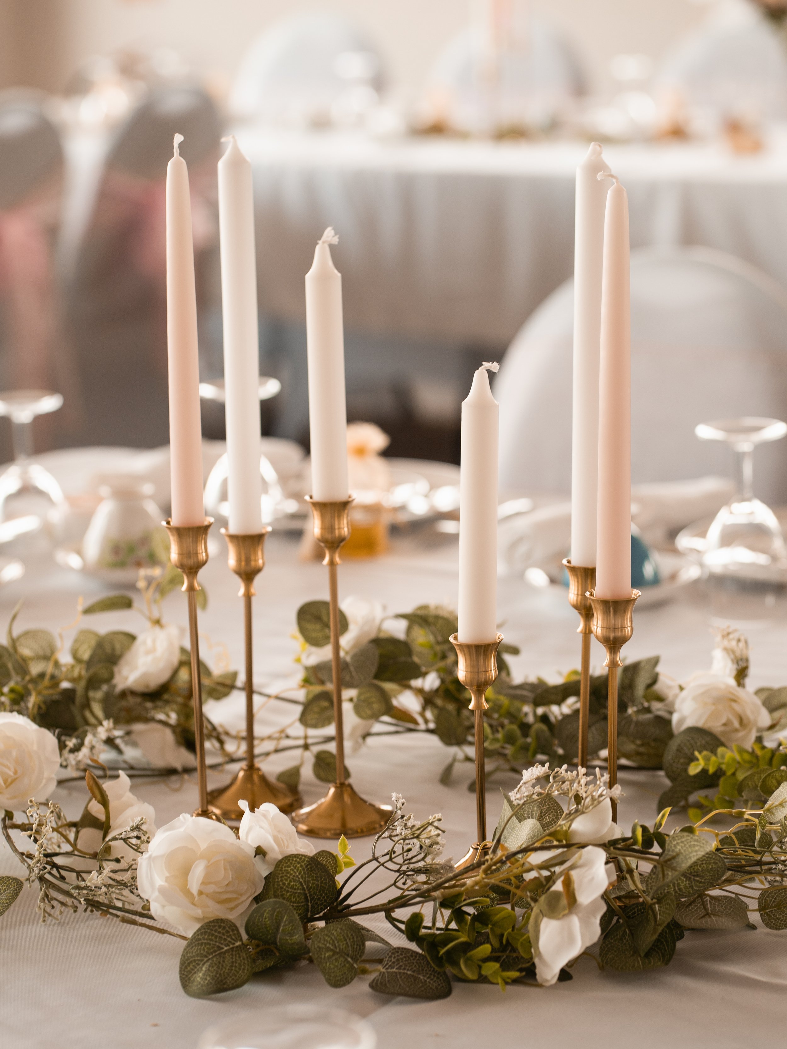 A table decorated with white candles in gold candle holders, surrounded by white flowers and greenery, likely for a wedding or special event.