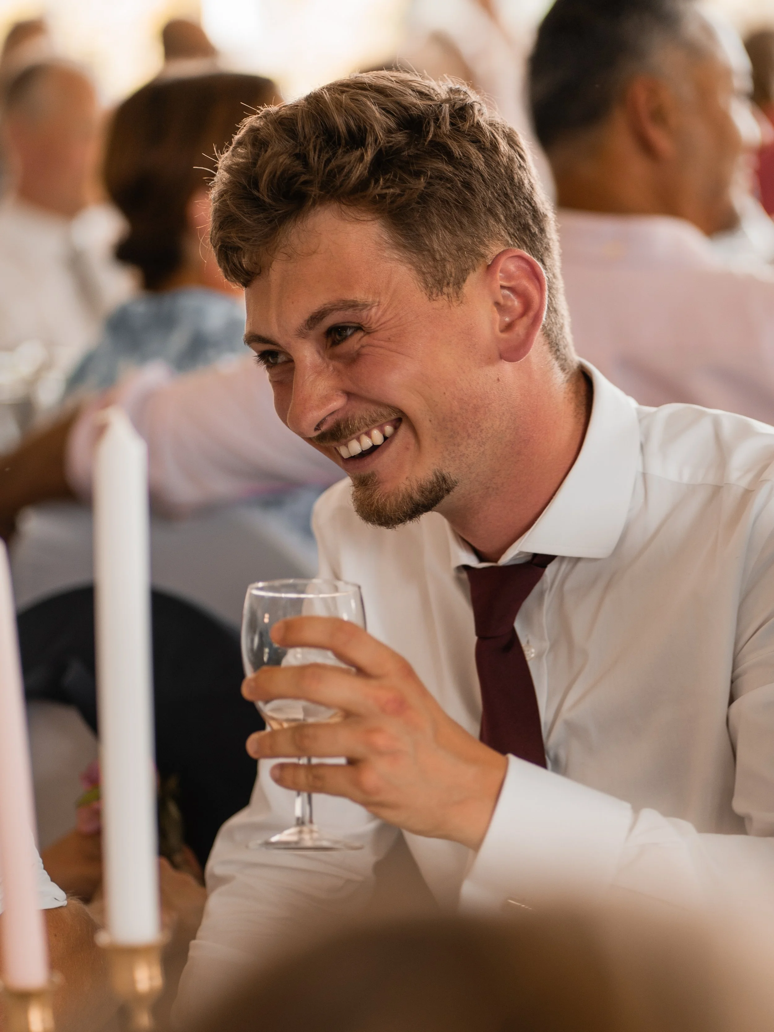 A man with short, curly hair and a trimmed beard, wearing a white shirt and a maroon tie, smiling and holding a glass of white wine at a social gathering or wedding reception.