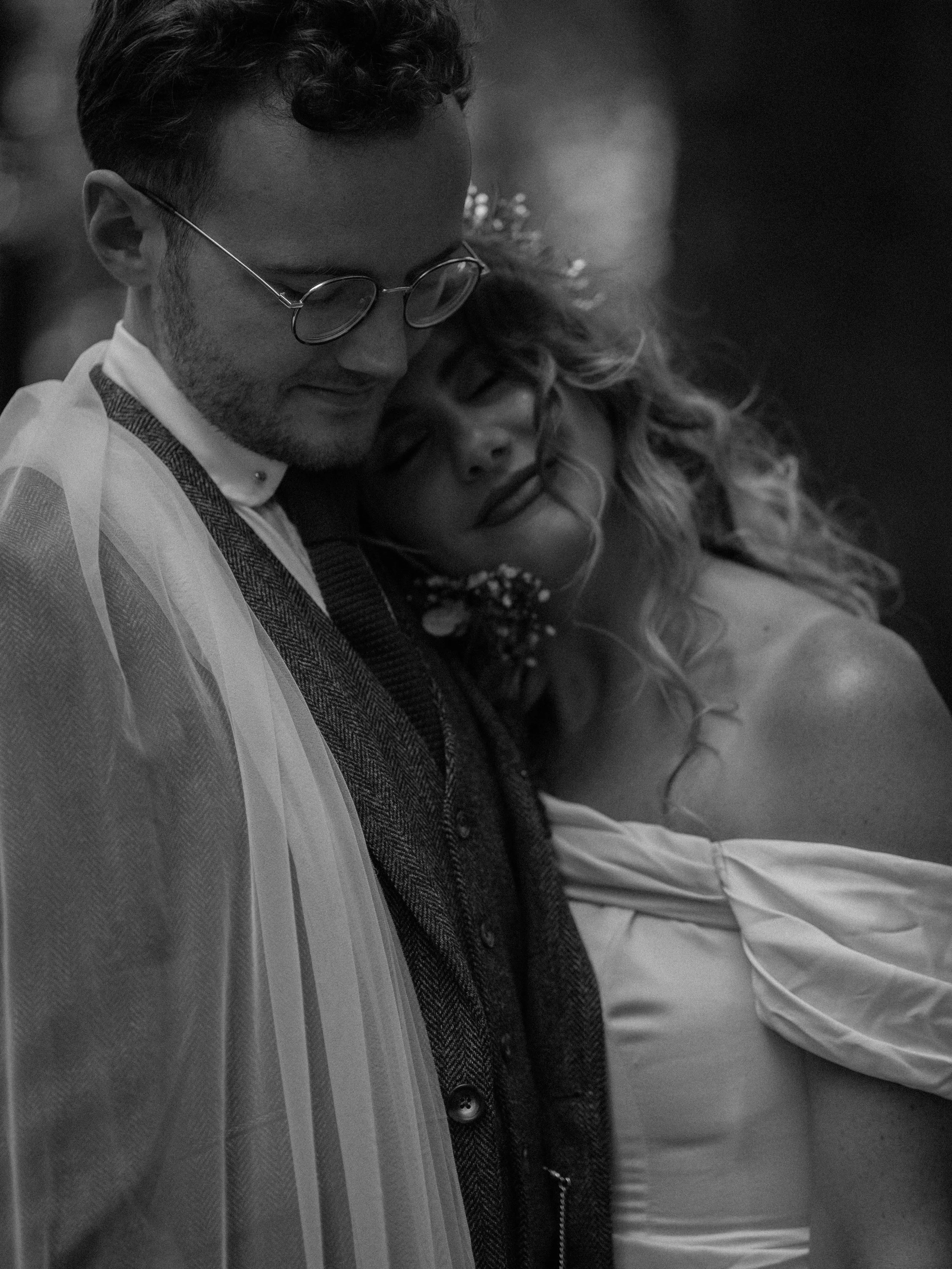 A black-and-white photo of a man and woman in a close embrace, with the woman resting her head on the man’s chest. The man is wearing glasses and a layered outfit, and the woman has curly hair and closed eyes.