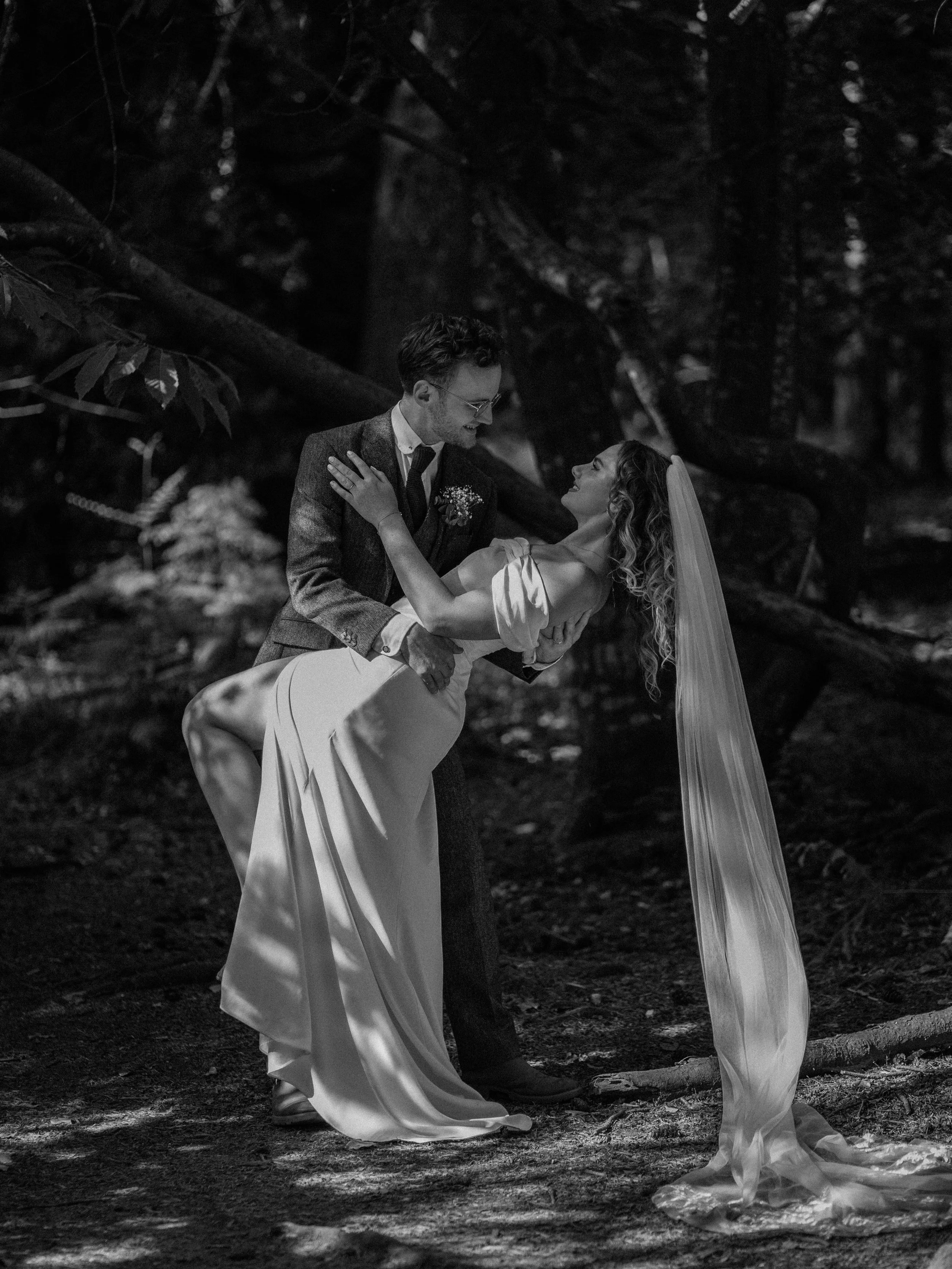 A black-and-white photo of a man in a suit and a woman in a wedding dress dancing outdoors under a tree in a wooded area.