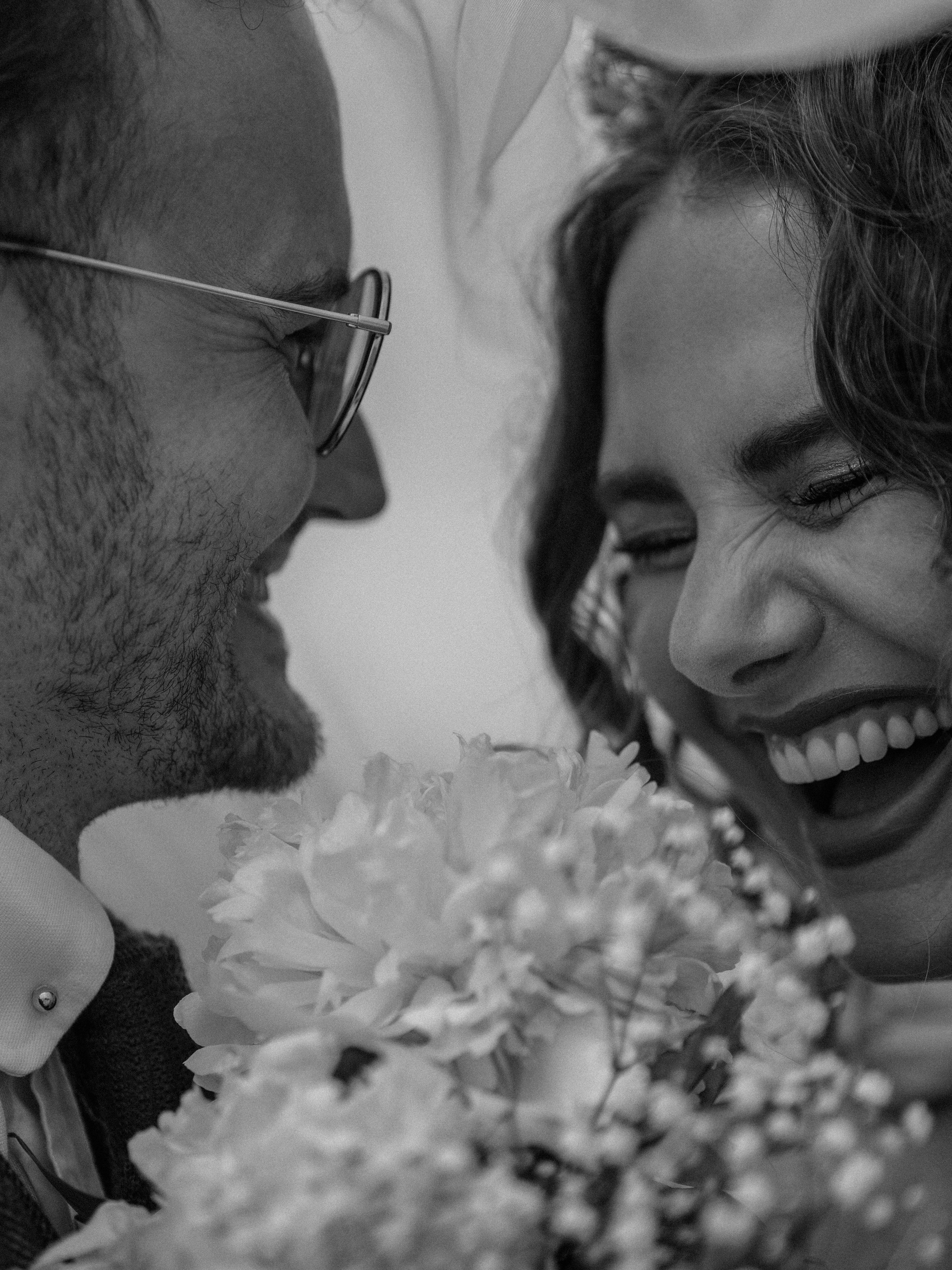A close-up black and white photo of a smiling woman with curly hair and a man with glasses, both with their foreheads touching, surrounded by flowers.