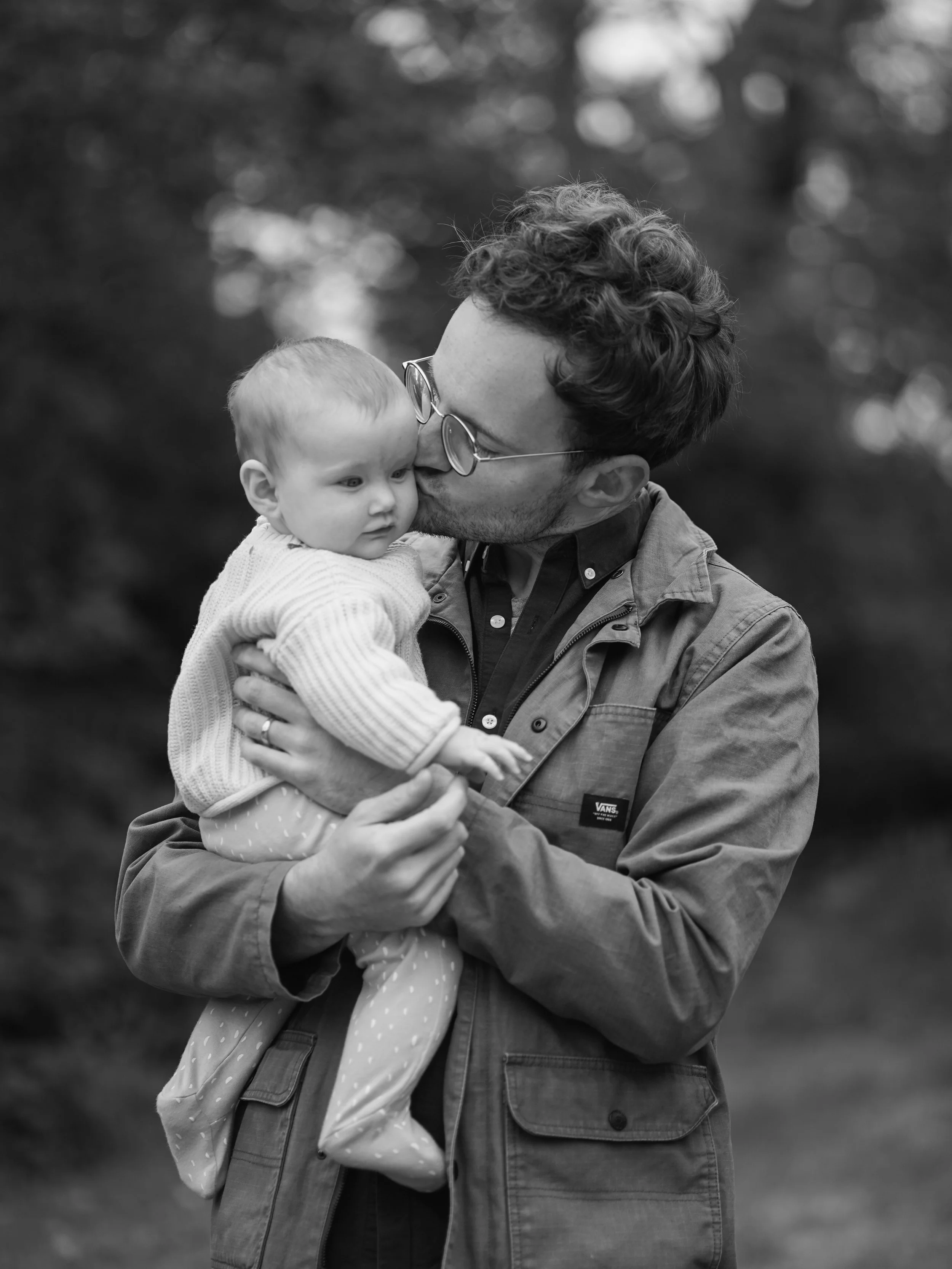 A man with glasses and curly hair holding a young girl, kissing her on the cheek outdoors with blurred trees in the background.