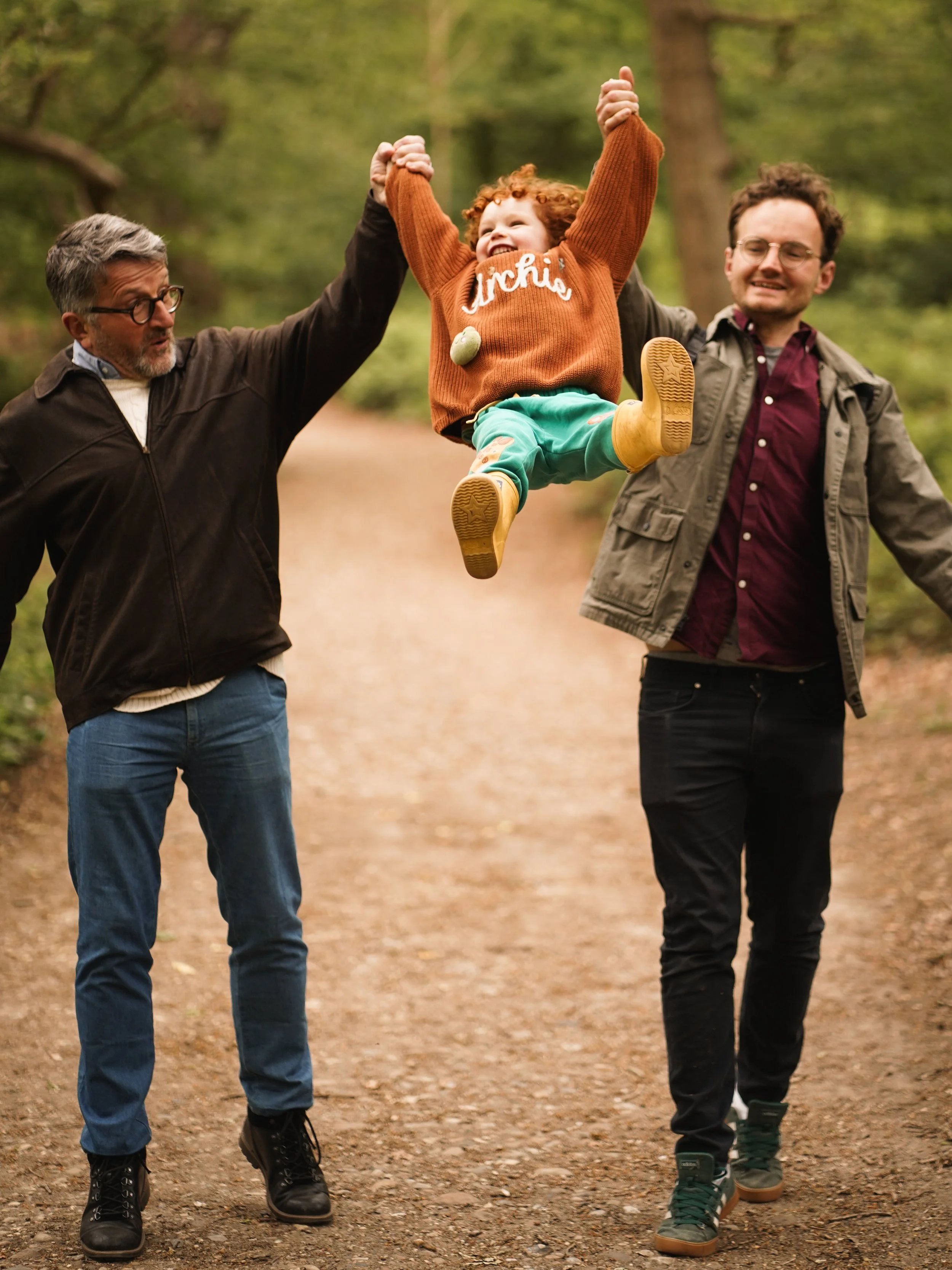 Two men walking on a forest trail while carrying a smiling child between them, holding her hands up in the air.