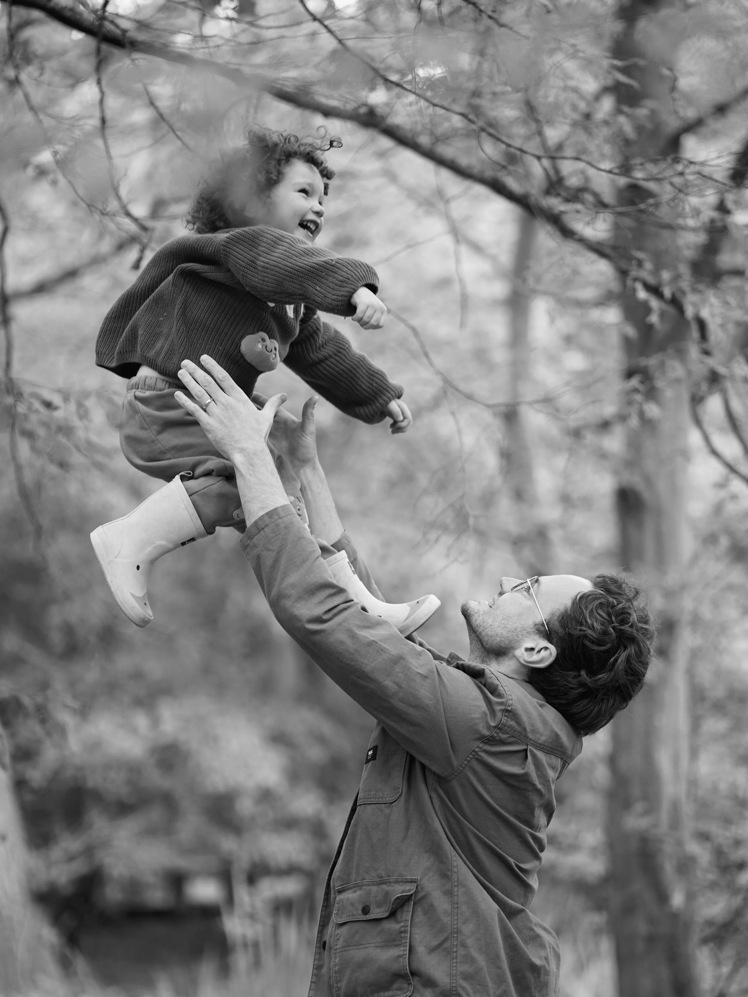 A man with glasses lifting a young girl into the air. The girl is smiling and pointing, with a background of trees and foliage.