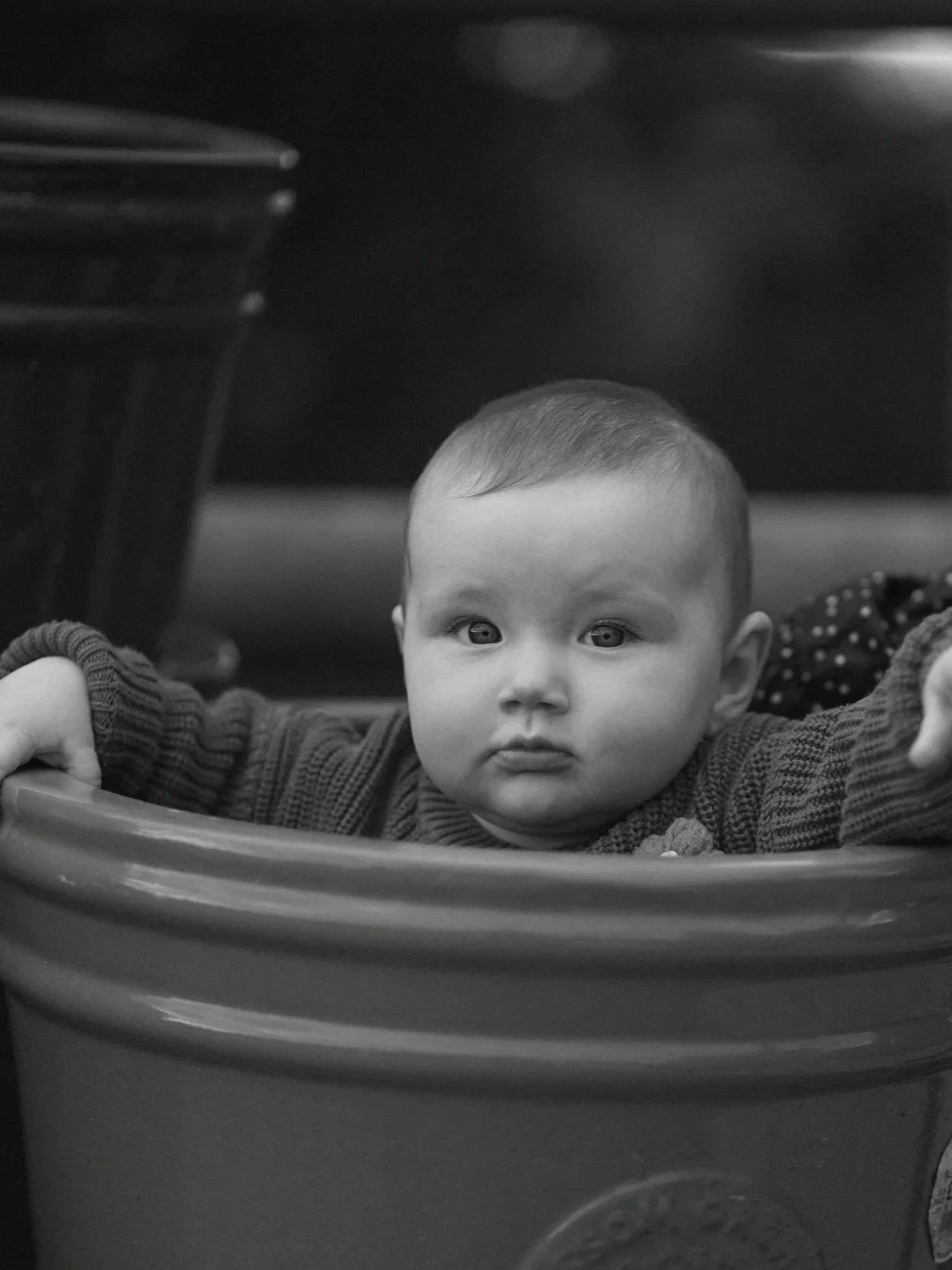 Black and white photo of a young child with short hair, wide eyes, and a neutral expression, lying in a plastic container or seat, with arms outstretched, in a room with a table in the background.