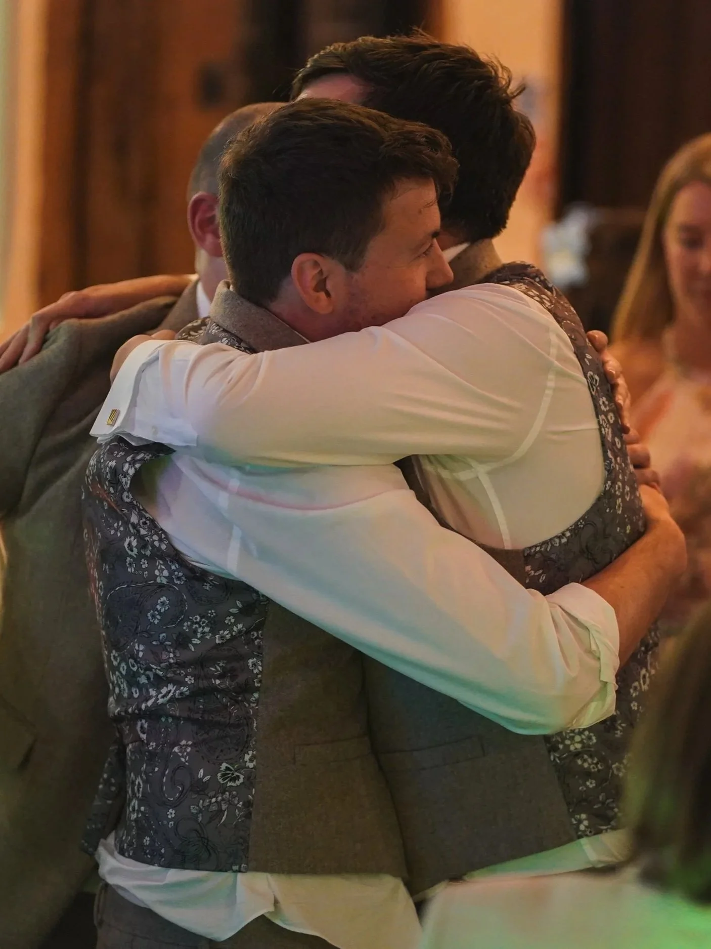 Two men hugging each other warmly at a social gathering, with other people visible in the background.