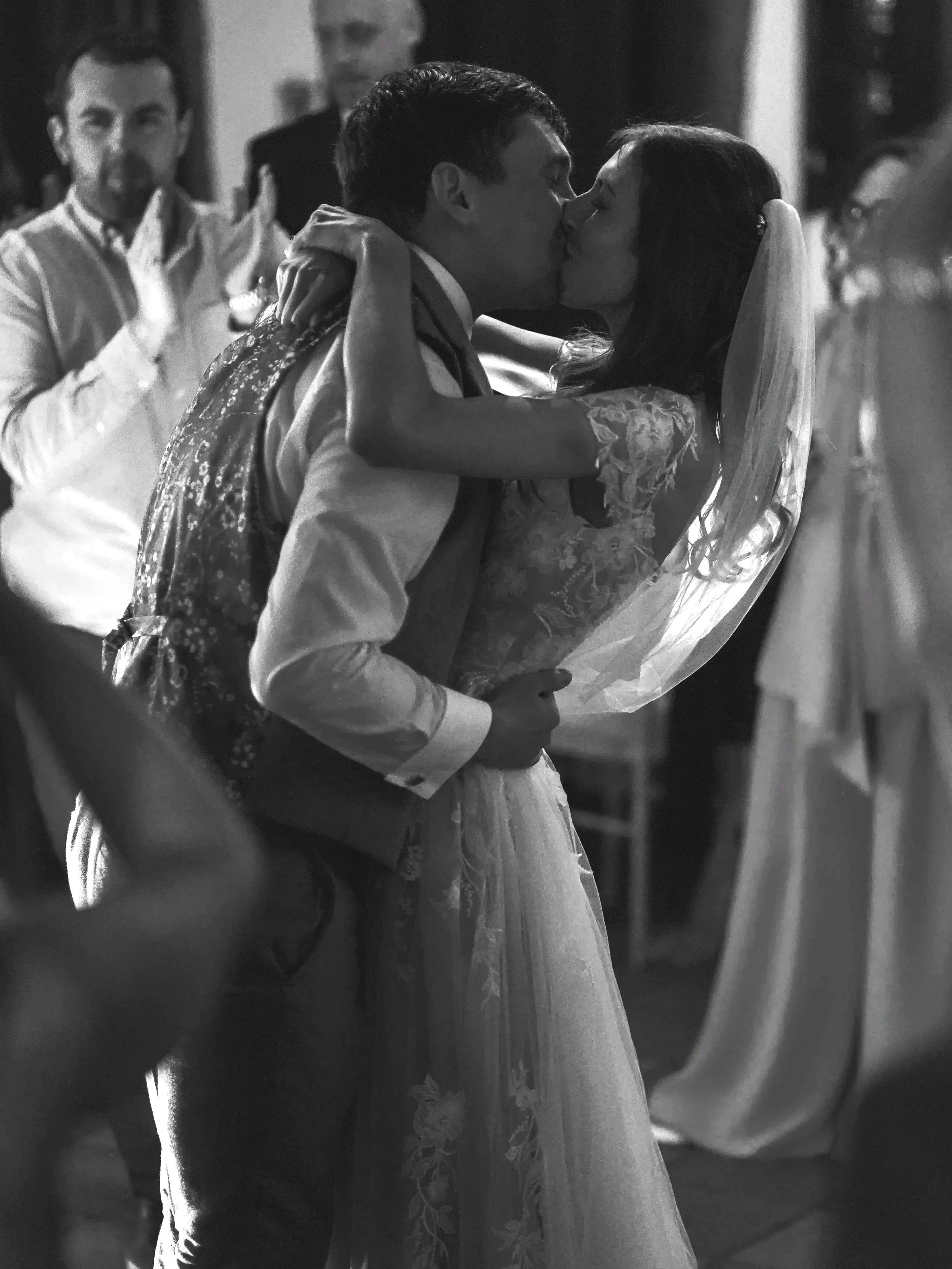 A black and white photo of a couple dancing and kissing at a formal event, with a man clapping in the background and other guests visible.
