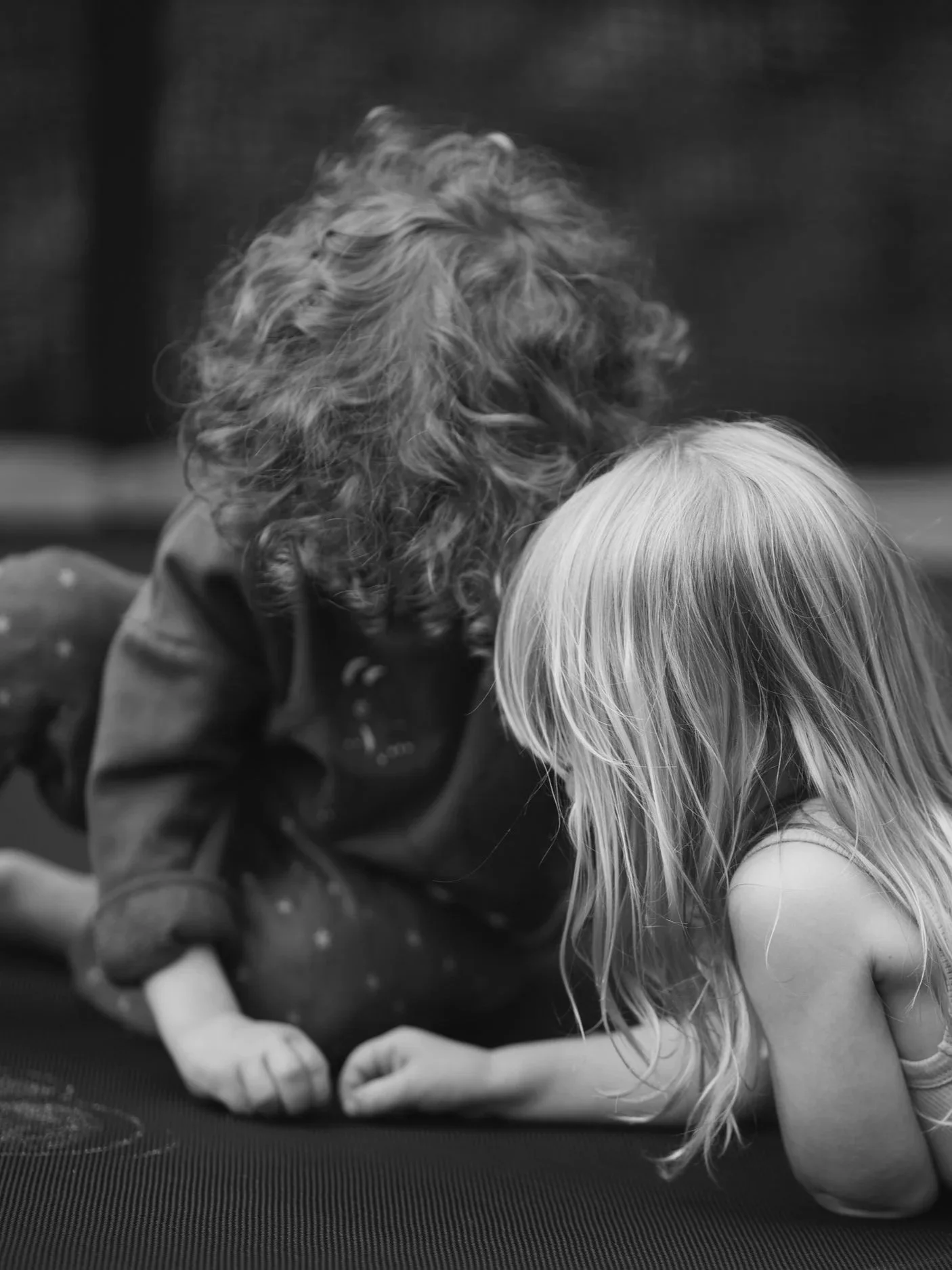 Two children, a girl with long blonde hair and a girl with curly hair, are sitting close together, looking downward, possibly engaged in an activity.